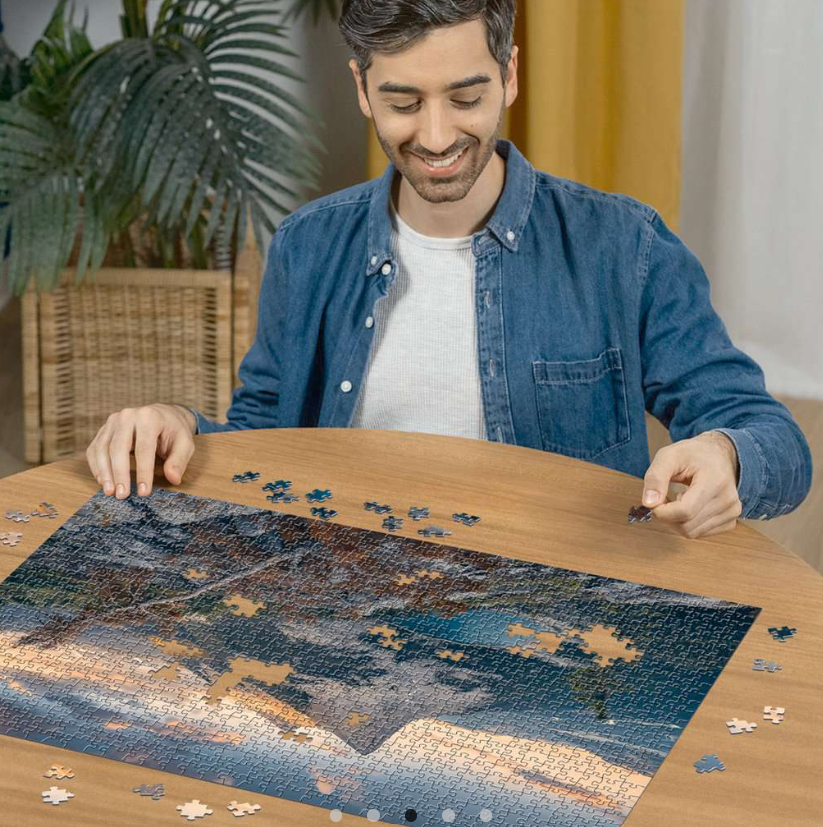 Ein Mann setzt ein Puzzle auf einem Holztisch zusammen. Das Puzzle zeigt eine Berglandschaft. Andere Puzzleteile sind verstreut.