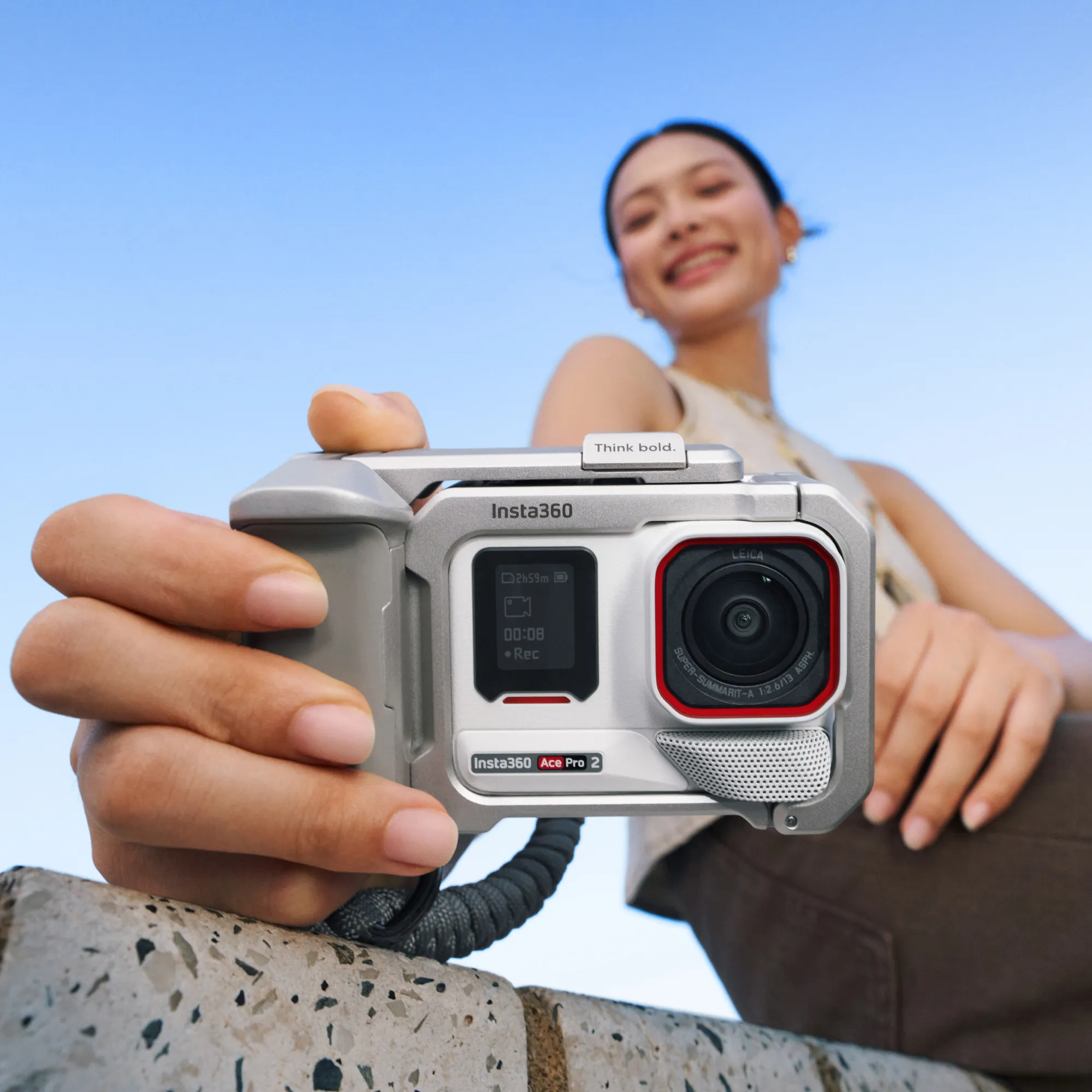 Eine Frau hält eine Insta360 Ace Pro 2 Kamera. Die Kamera ist silber und schwarz, mit einem roten Ring um das Objektiv. Blauer Himmel im Hintergrund.