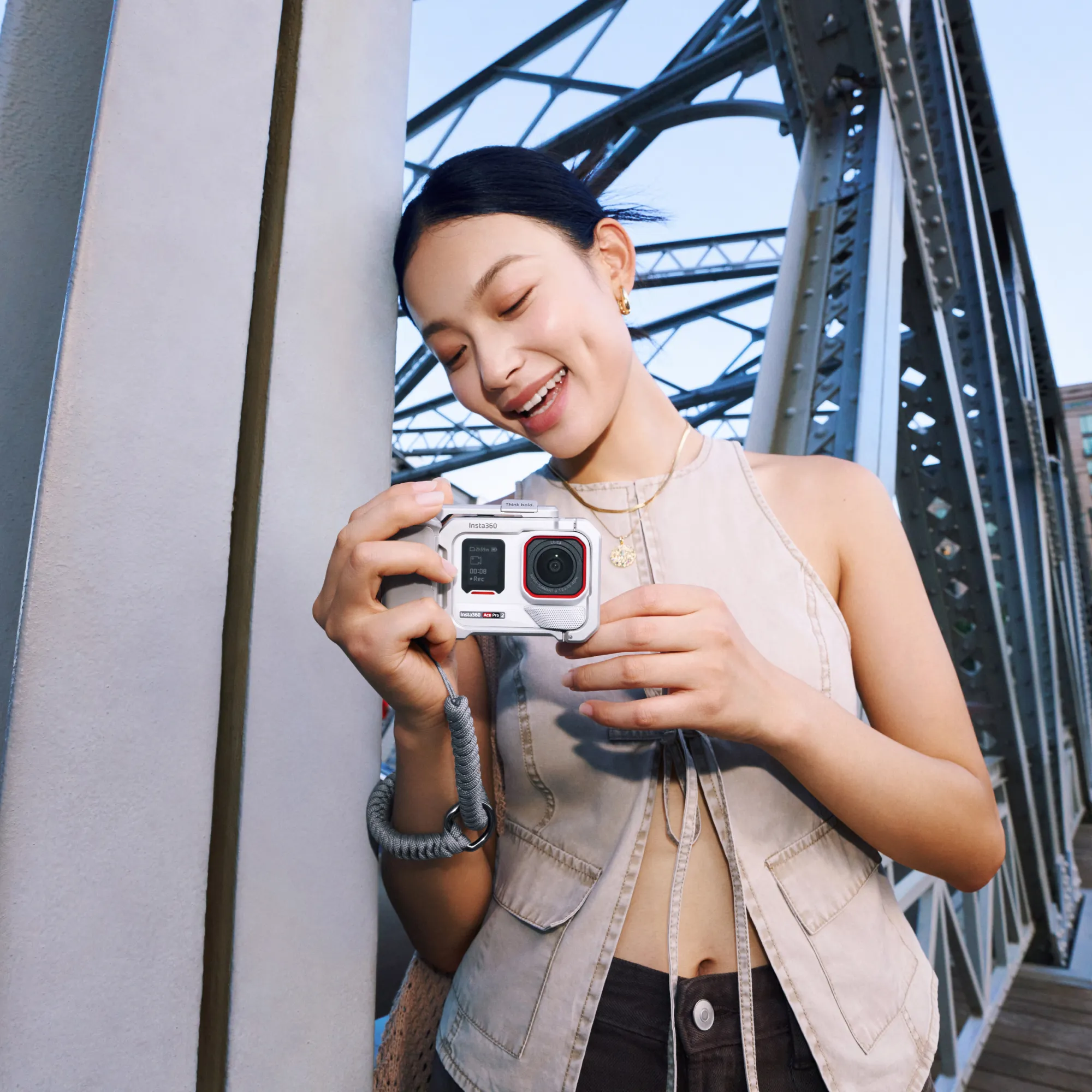 Frau lächelt, hält Kamera auf Brücke. Kamera ist Insta360 Ace Pro. Hintergrund zeigt Brückenstruktur und Himmel.