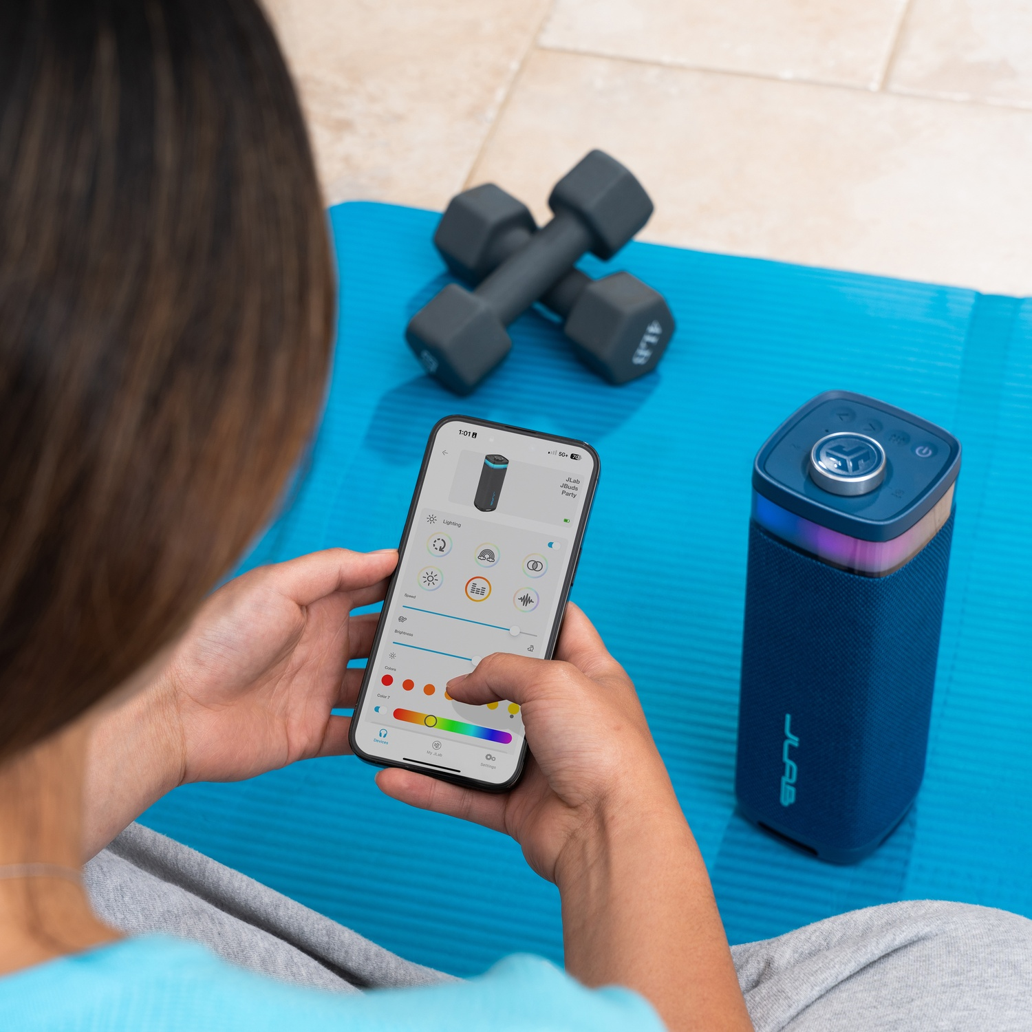 A person is using a phone to control a blue speaker. Dumbbells and a blue mat are in the background.