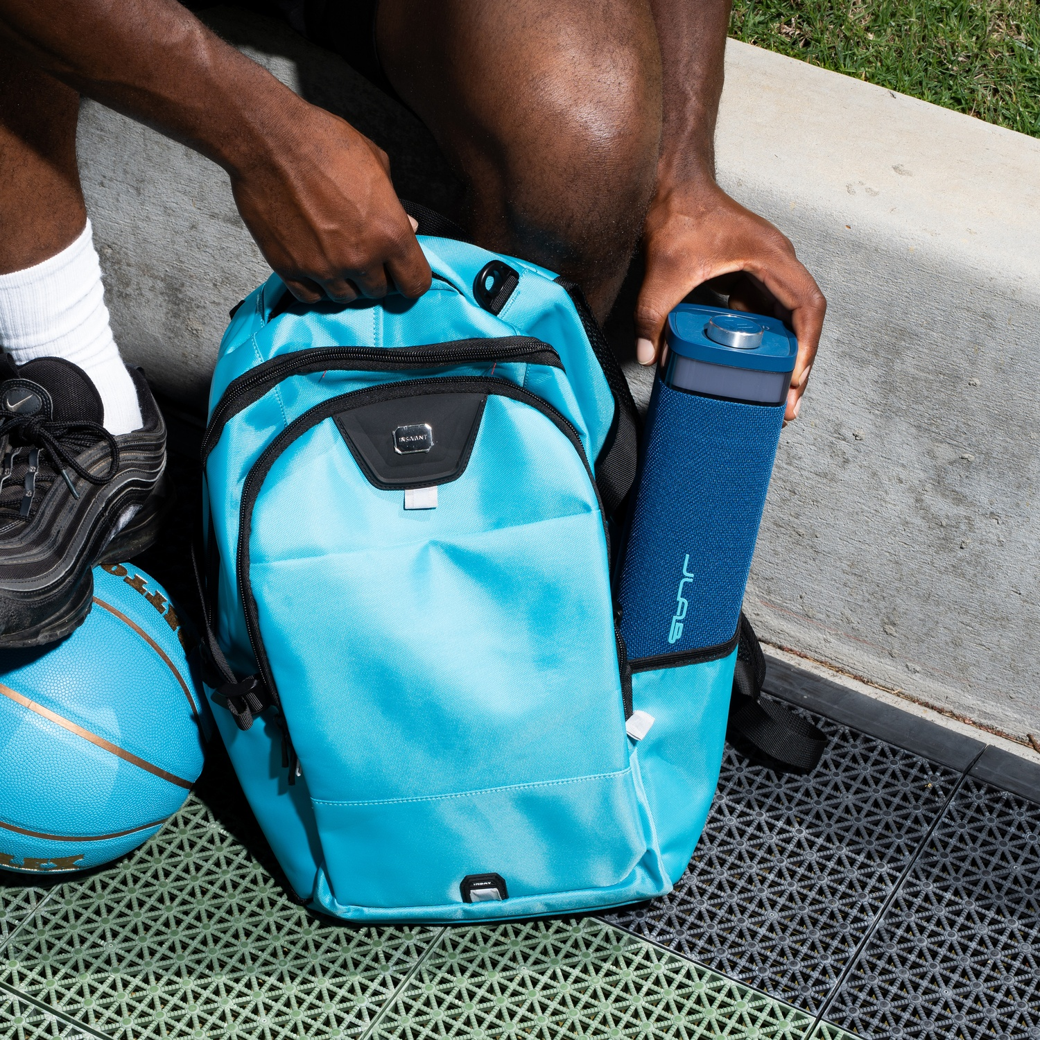 A person is holding a blue speaker from a blue backpack. Basketball, shoe, and concrete are also visible in the image.