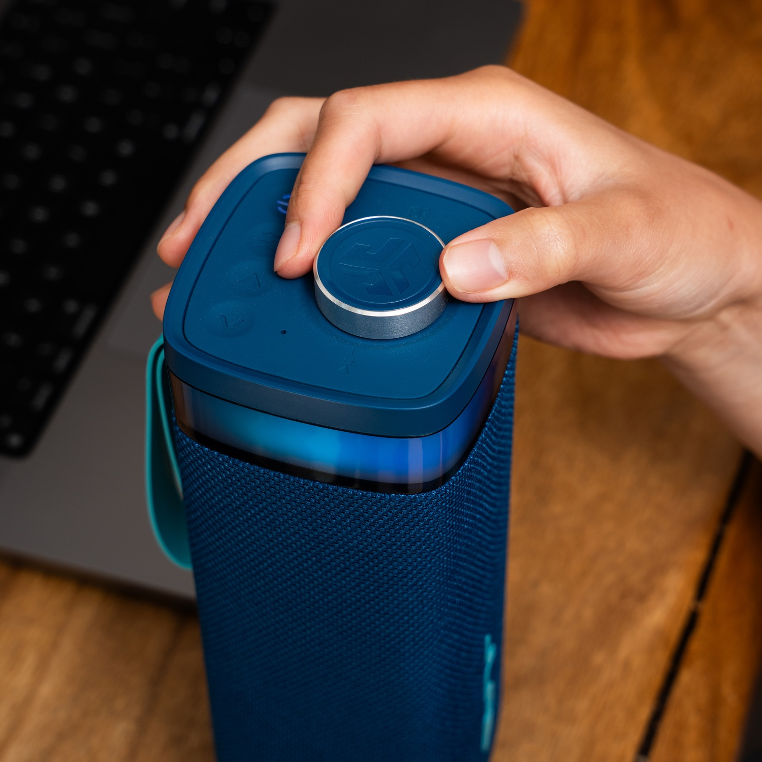 A hand adjusts the volume on a blue speaker. The speaker sits on a wooden surface next to a laptop.