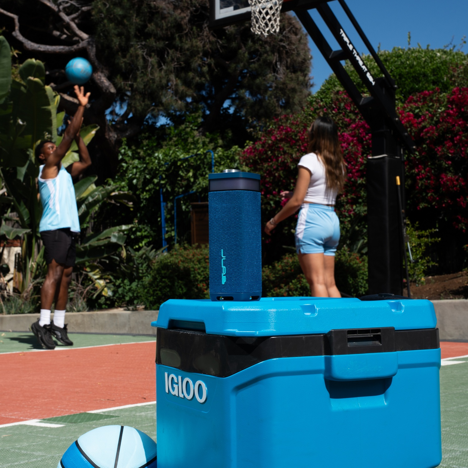 A man shoots a blue basketball. A woman stands near a speaker on a cooler. A ball is on the court.