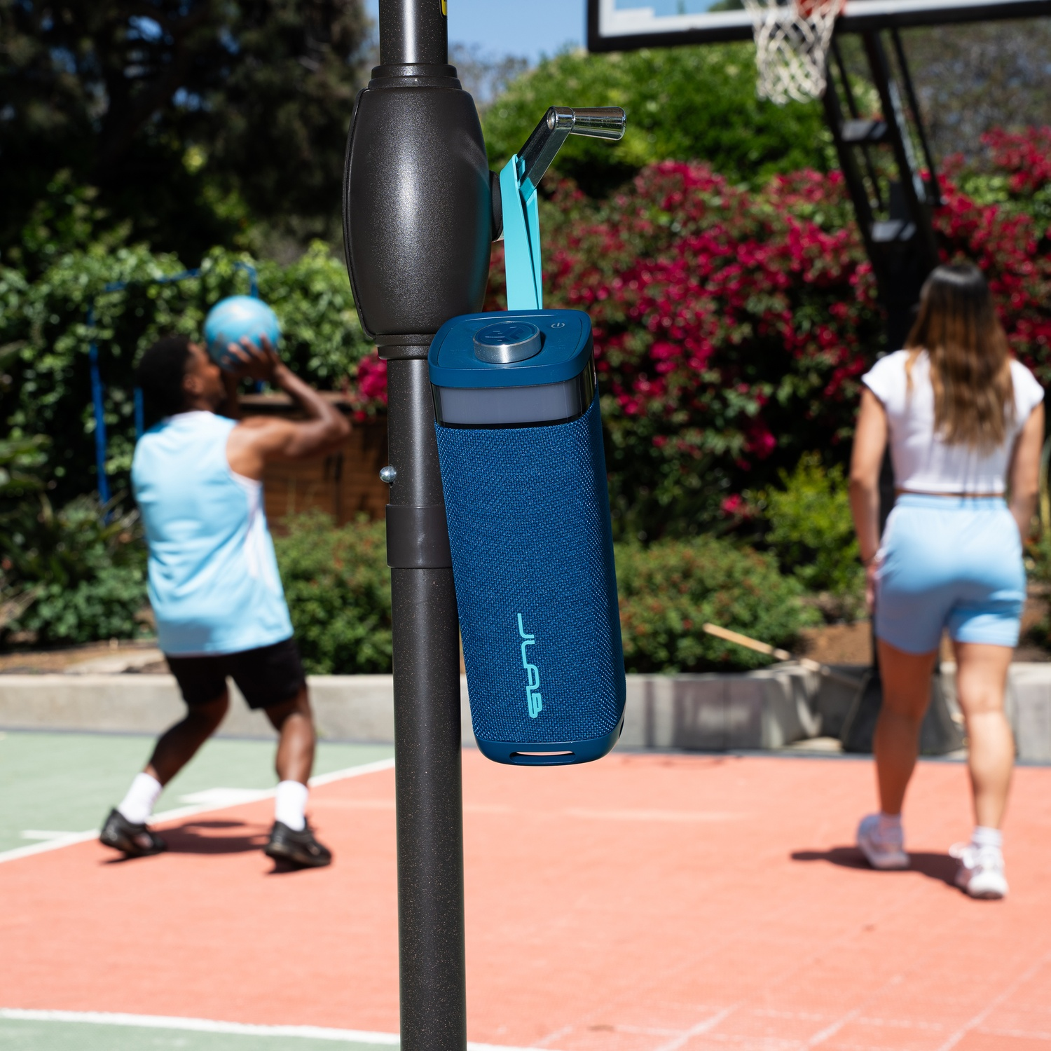 A blue speaker hangs from a basketball hoop pole. A person throws a ball, another walks away. A basketball court is in the background.