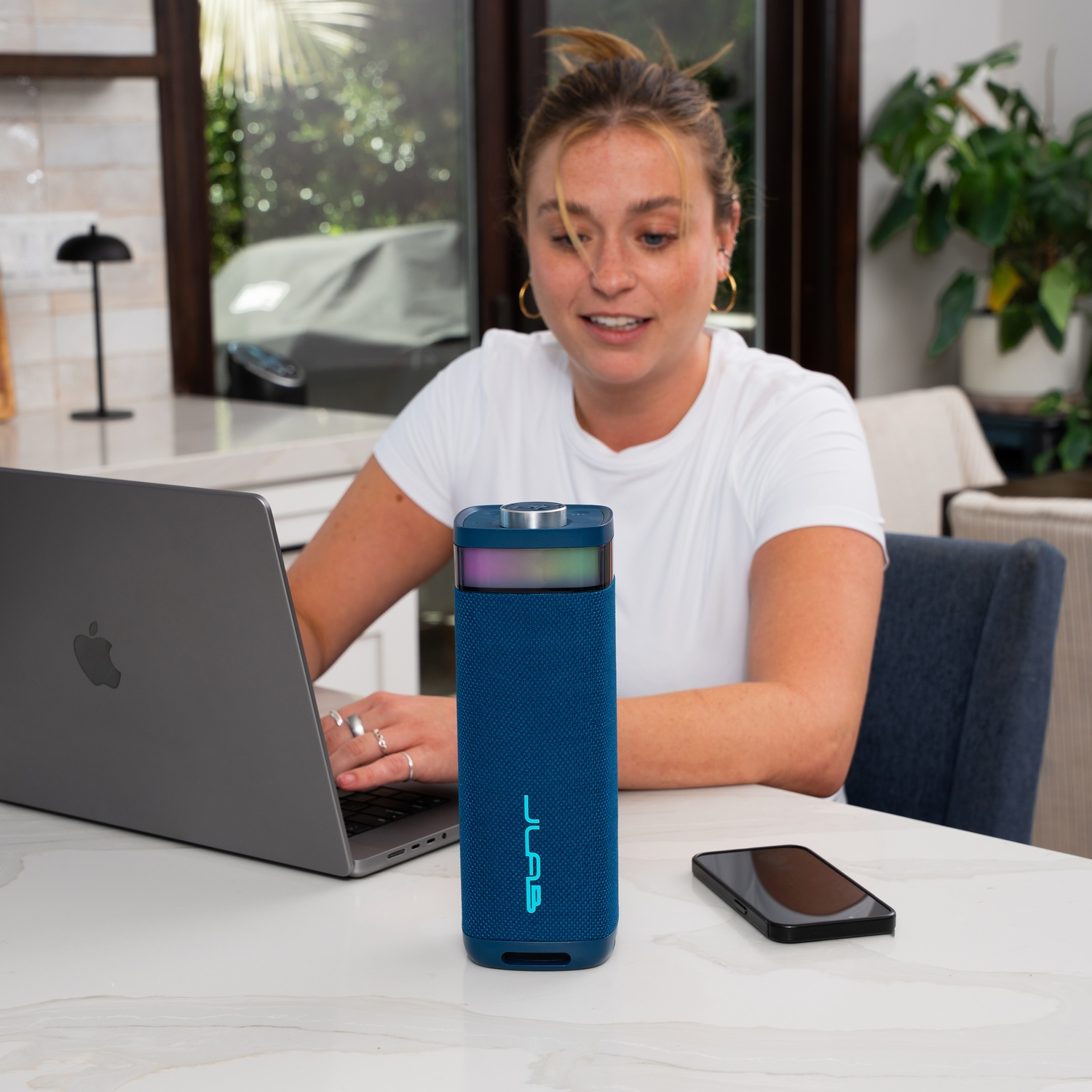 Woman uses a laptop, blue speaker, phone on table. Plants, window, and chair are in the background.
