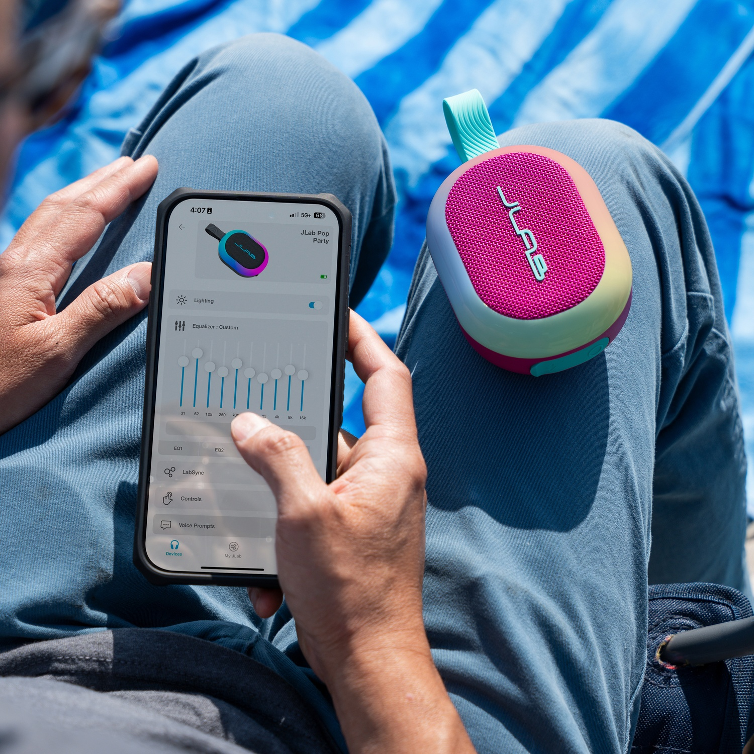 Une personne tient un smartphone et un haut-parleur JLab. Le téléphone affiche une interface d'application. Le haut-parleur est rose et coloré.