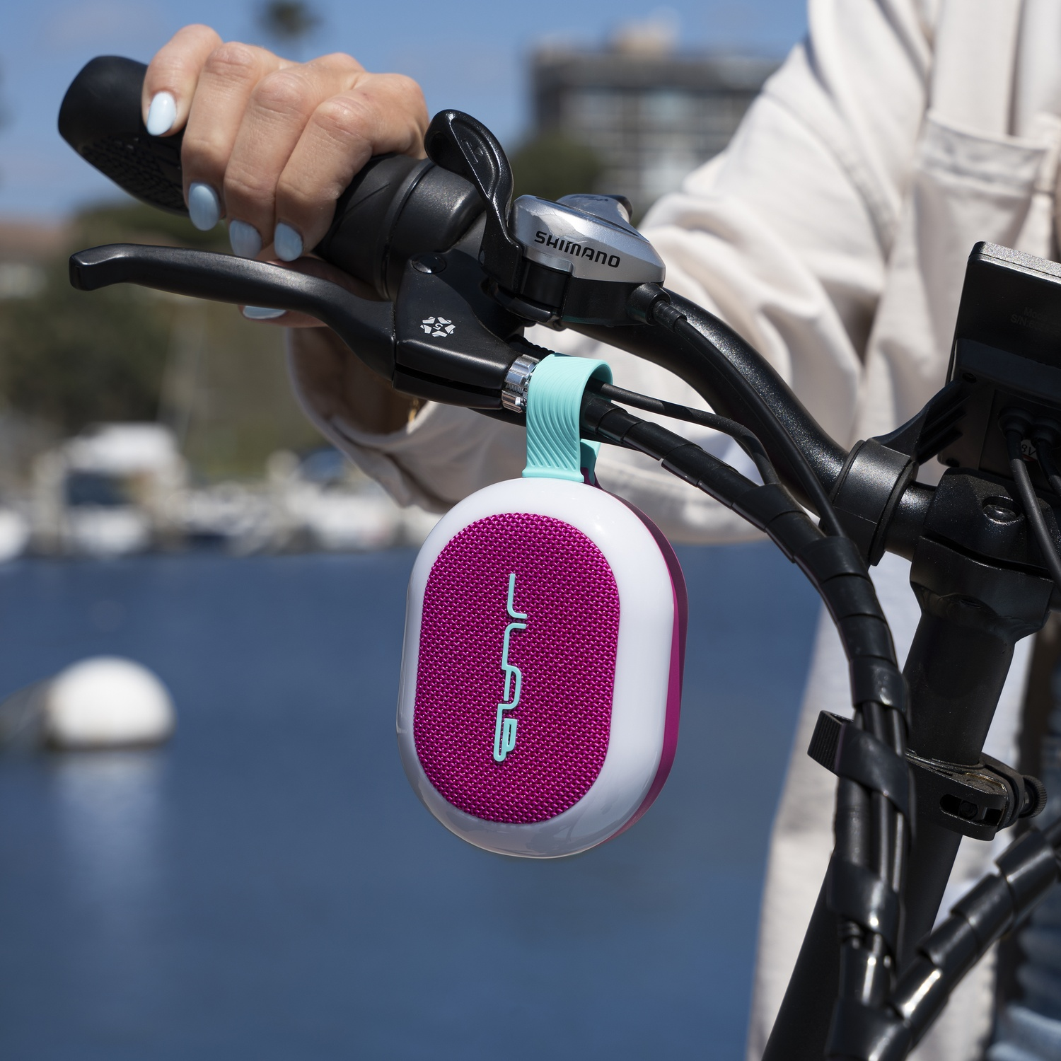 Une personne tient un guidon de vélo avec un haut-parleur attaché. Le haut-parleur est rose et blanc. Une mer bleue est en arrière-plan.
