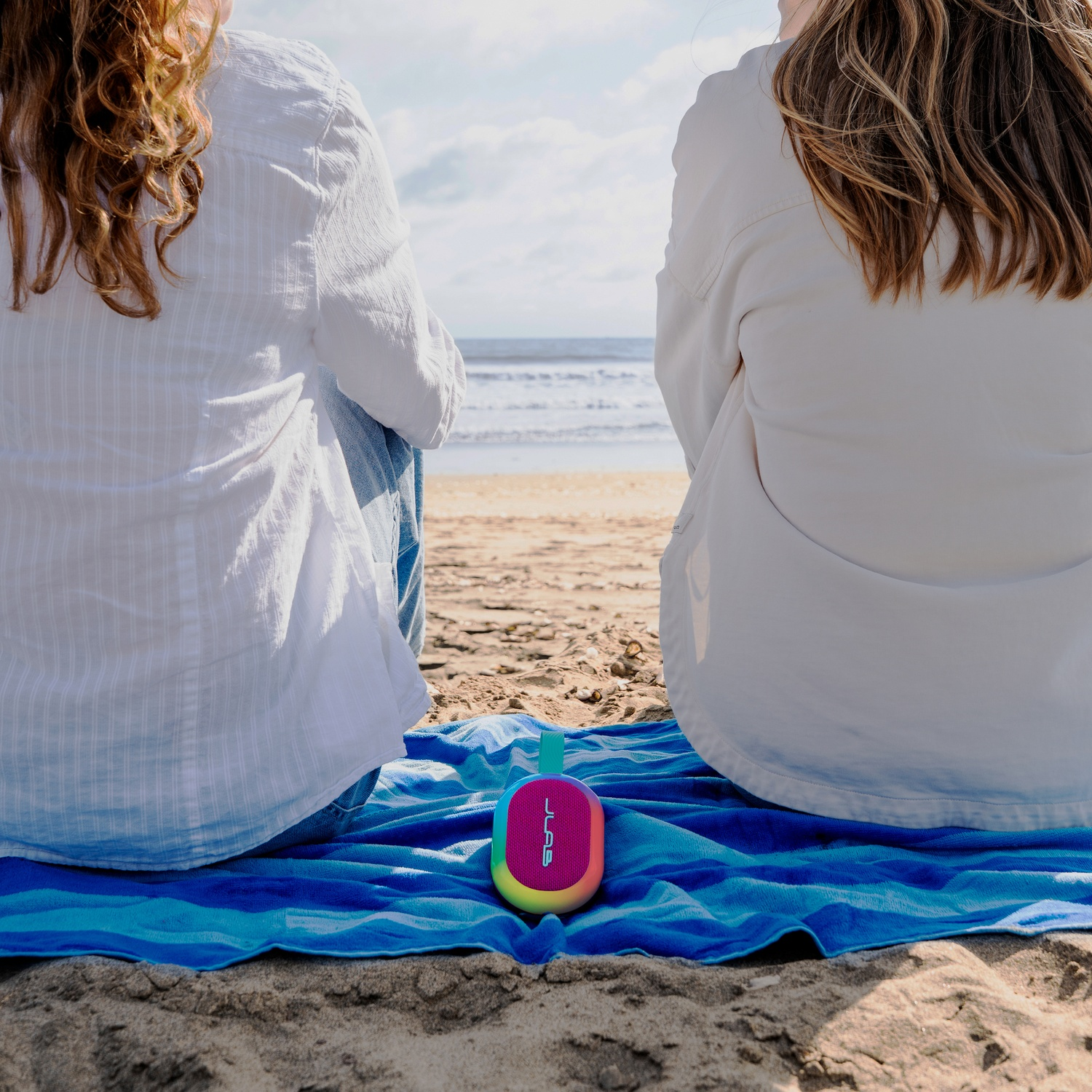 Deux femmes sont assises sur une plage avec un haut-parleur. Des vagues et le ciel sont en arrière-plan. Elles portent des chemises blanches.