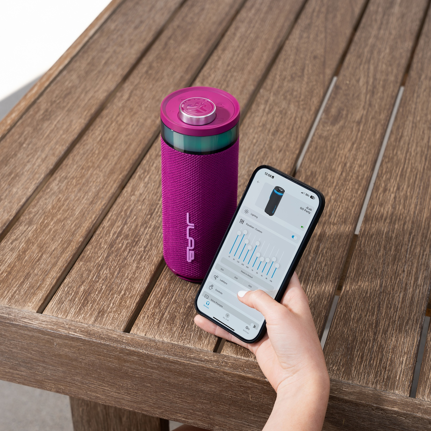 Een paarse luidspreker en een telefoon met een app-interface, beide op een houten tafel. De telefoon wordt door een persoon vastgehouden.