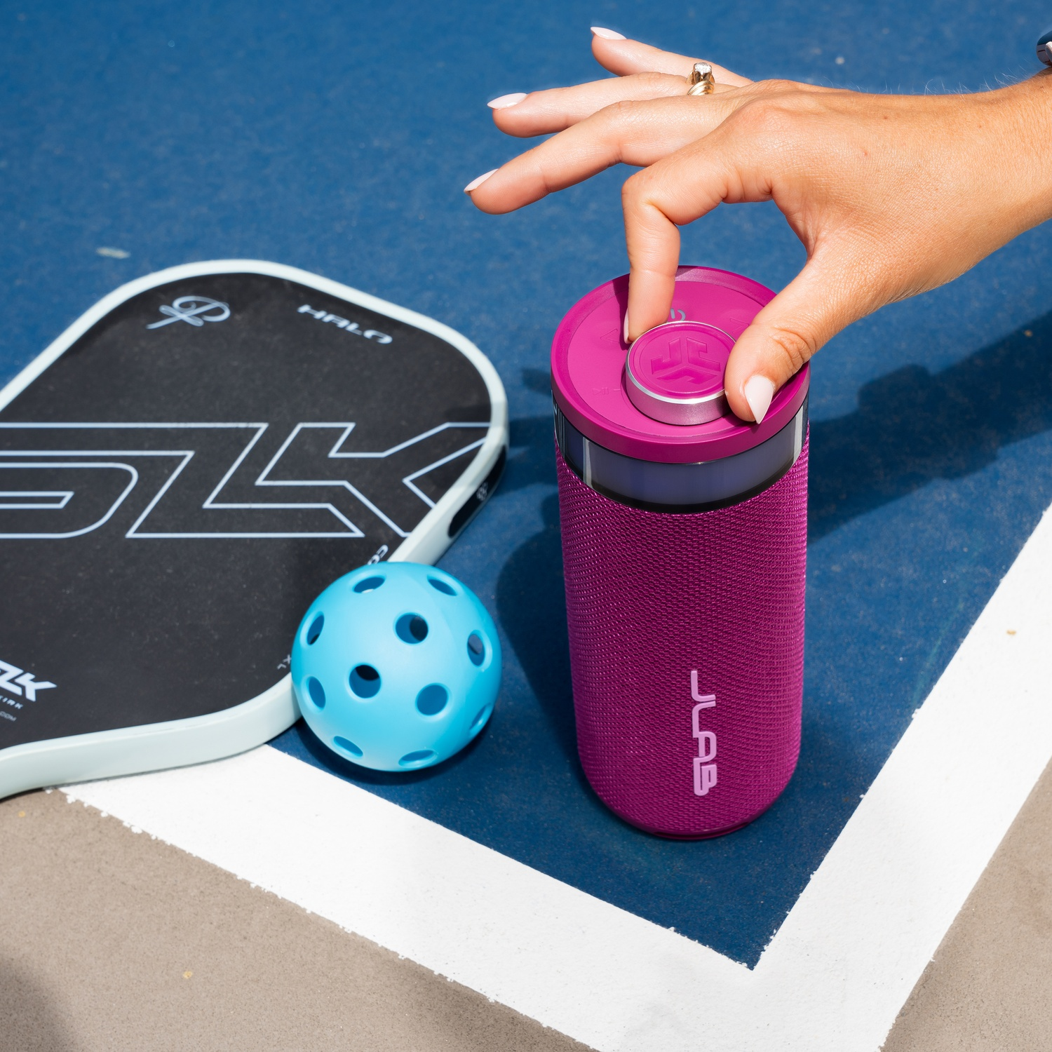 Een hand verstelt een roze luidspreker op een baan. Een pickleball racket en bal zijn in de buurt.