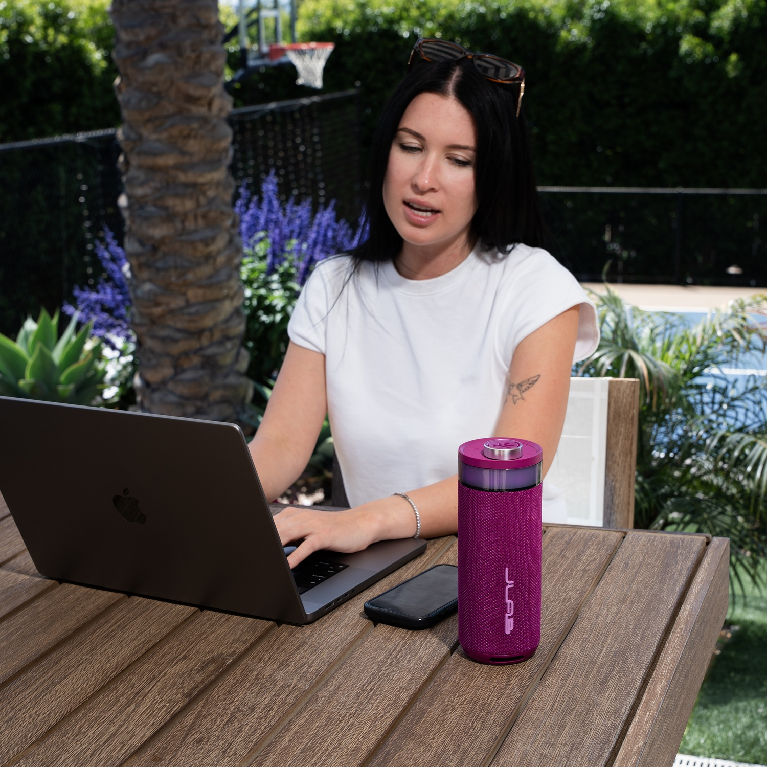 Vrouw met laptop buiten. Een paarse luidspreker en telefoon liggen op tafel. Zonnebril op haar donkere haar.