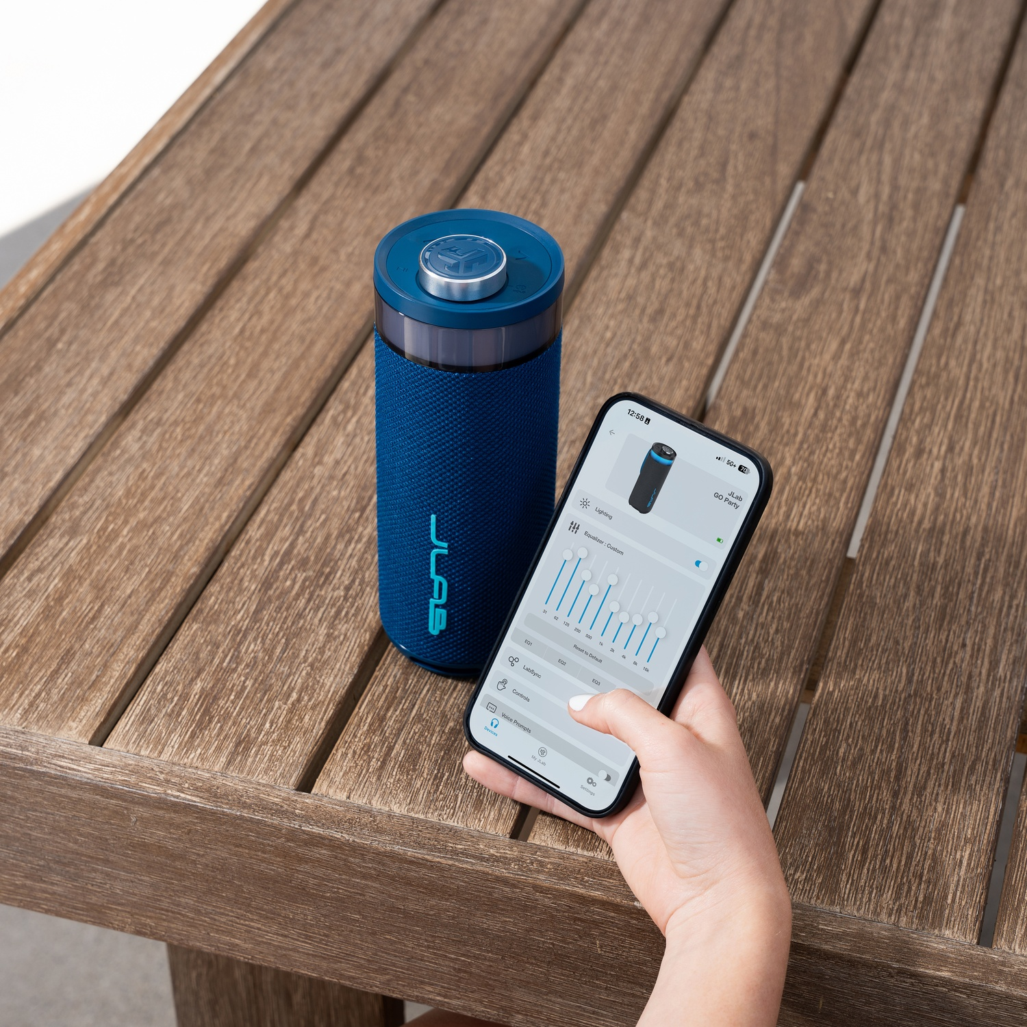Een blauwe luidspreker en een telefoon met een app-interface liggen op een houten tafel. De telefoon wordt door een persoon vastgehouden.