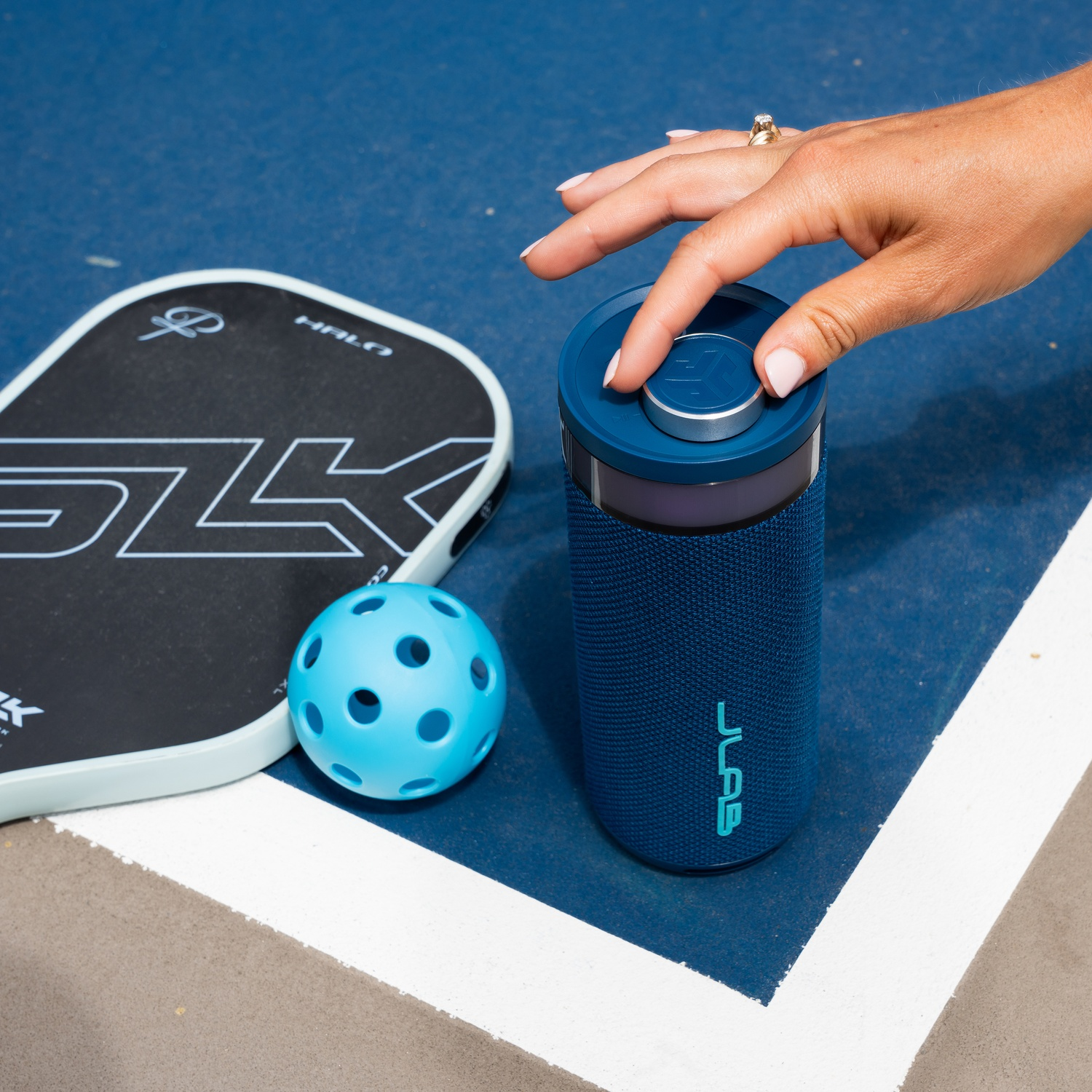 Een hand op een blauwe luidspreker, naast een pickleball-peddel en -bal op een blauwe baan.