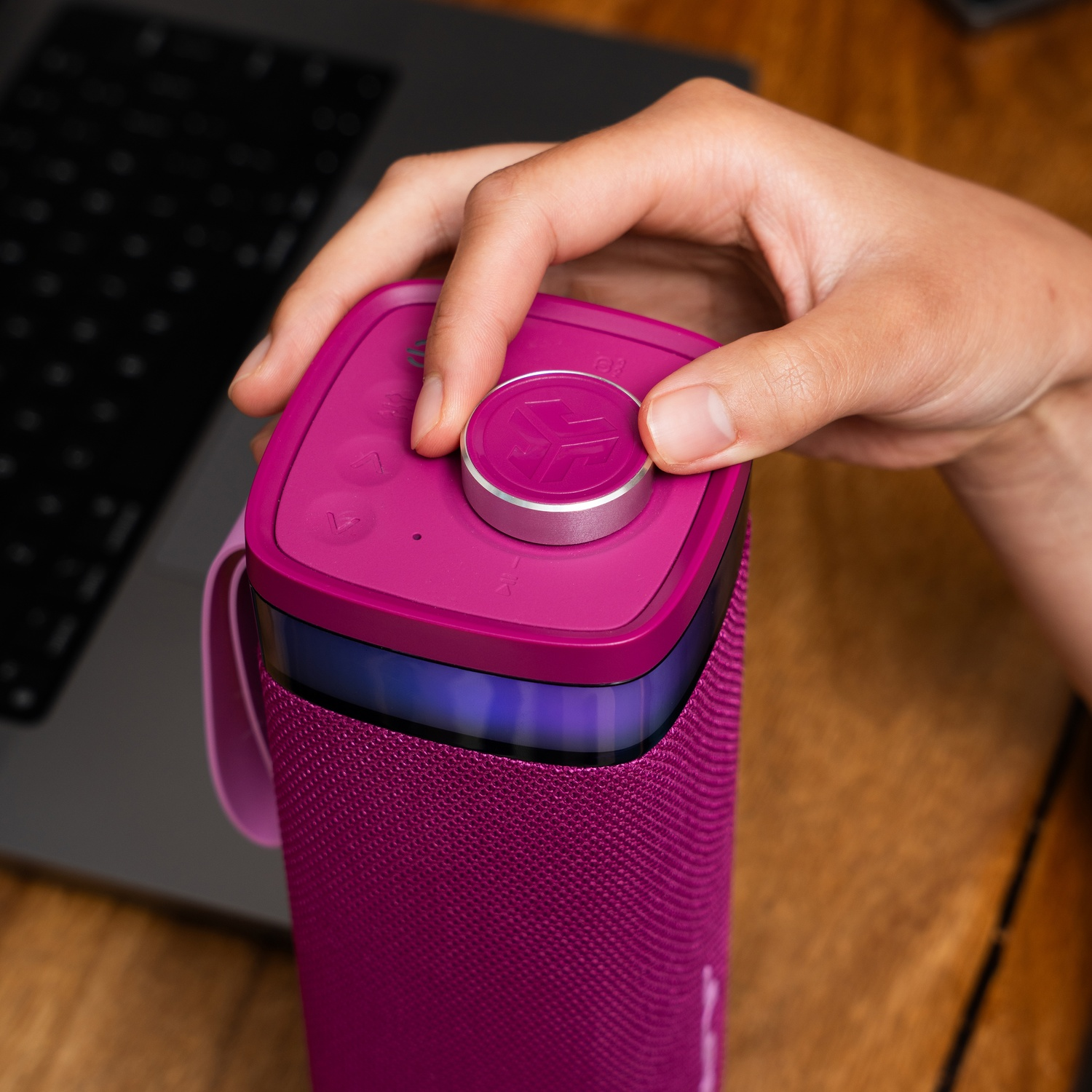 A hand adjusts the volume knob on a purple speaker. A laptop is in the background, sitting on a wooden desk.