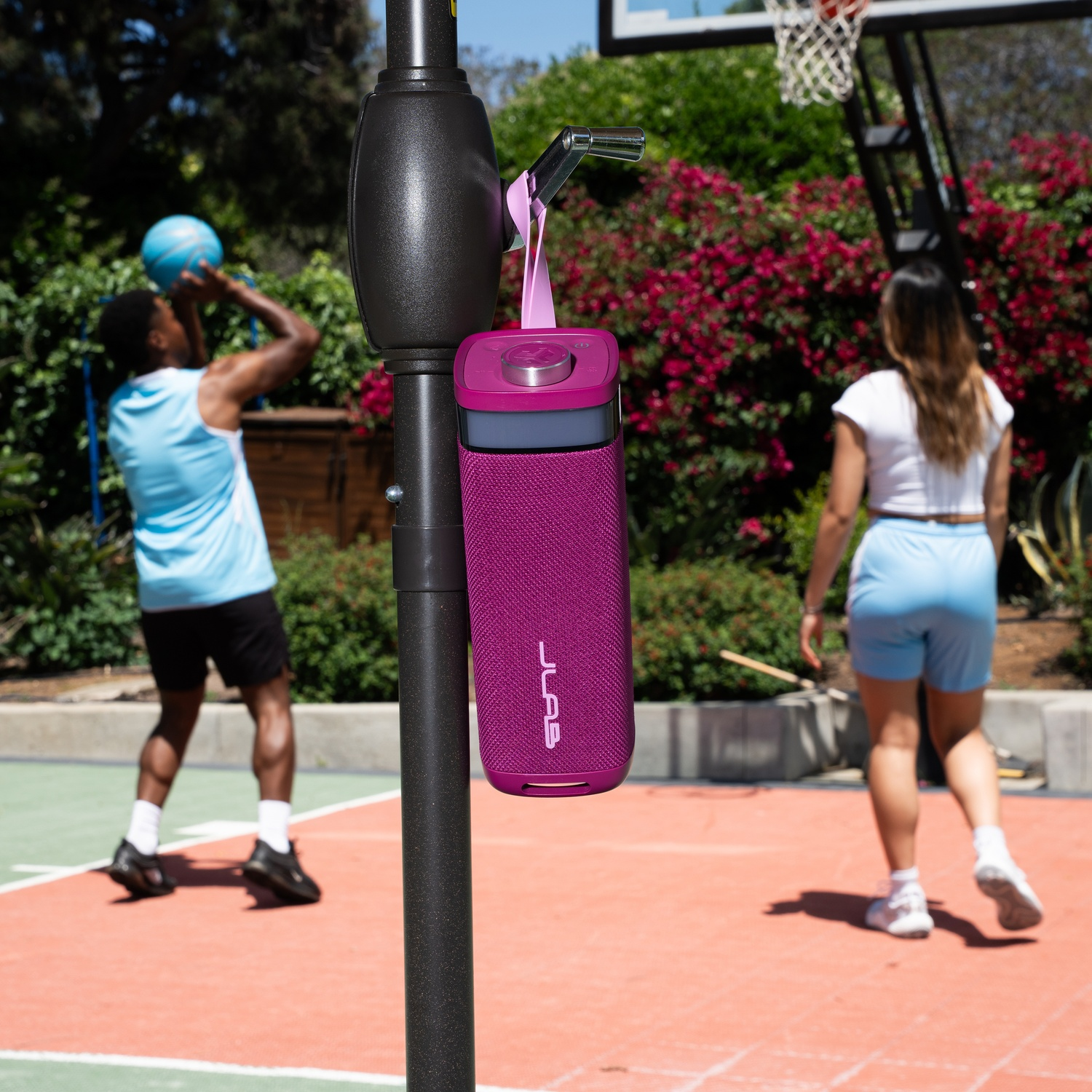 A purple speaker hangs on a basketball hoop. Two people play basketball. One shoots, the other walks away.