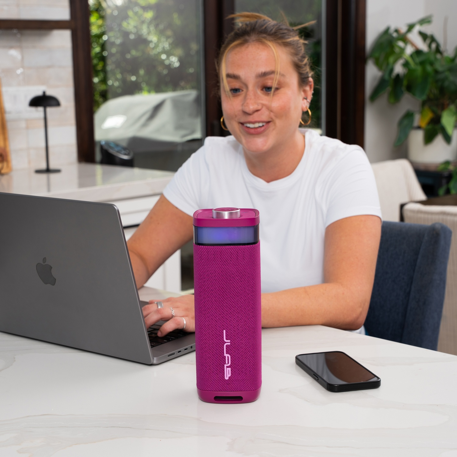Woman smiles, using laptop. A pink speaker and phone sit on the table. A window and plants are in the background.