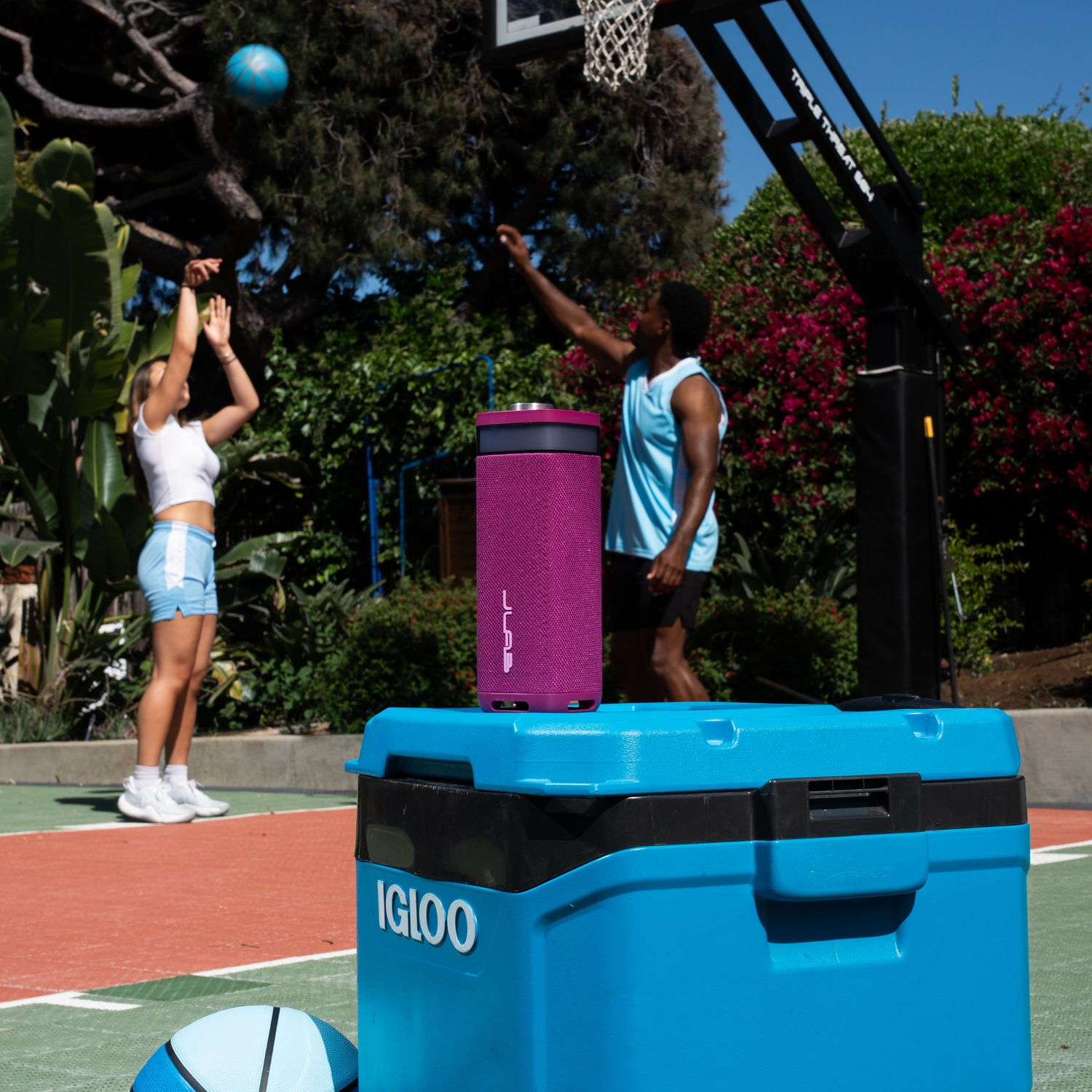 Two people playing basketball outdoors. A purple speaker sits on a blue cooler. A basketball in flight.