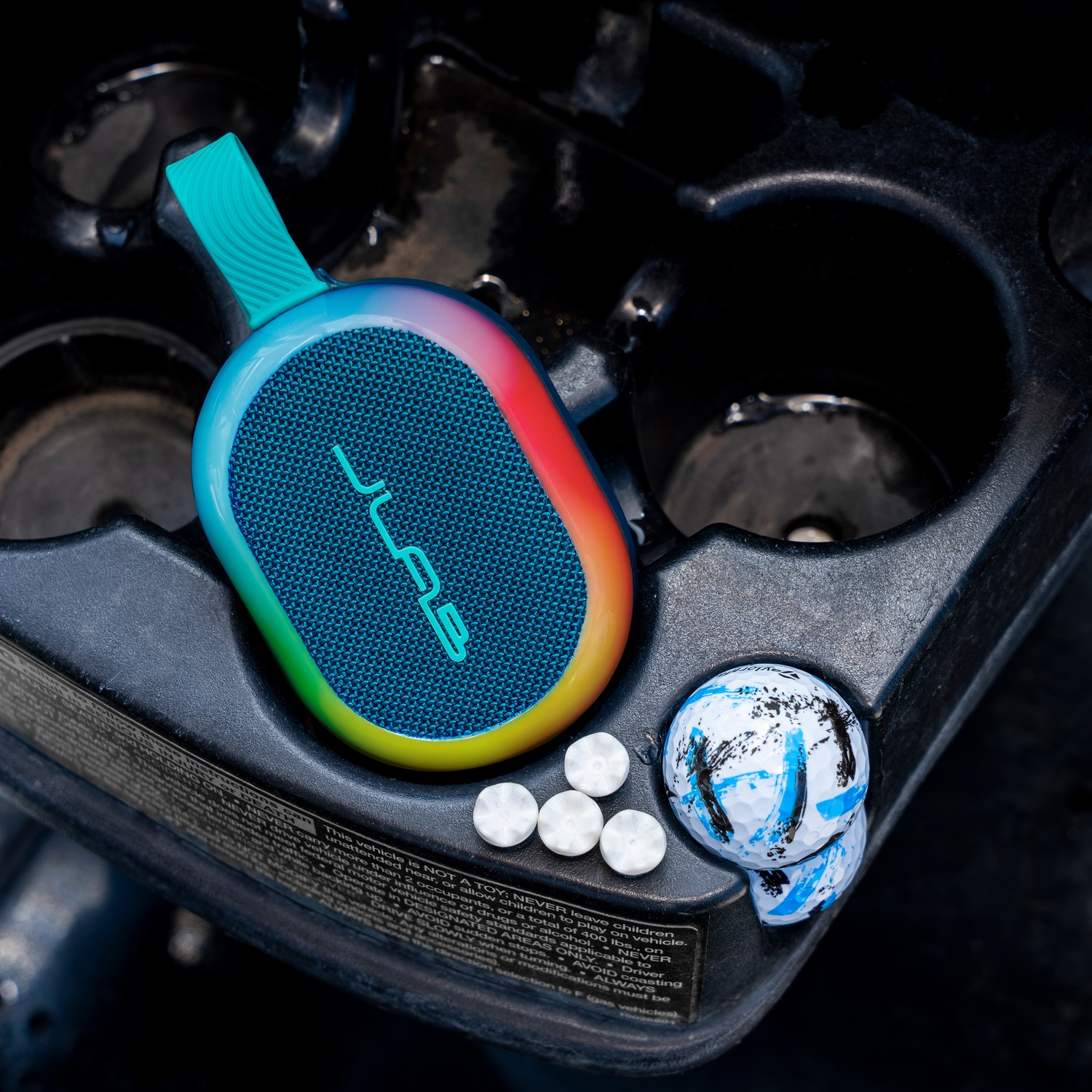 Een kleurrijke speaker, pillen en een golfbal in een bekerhouder in de auto. De speaker heeft het JLab-logo.