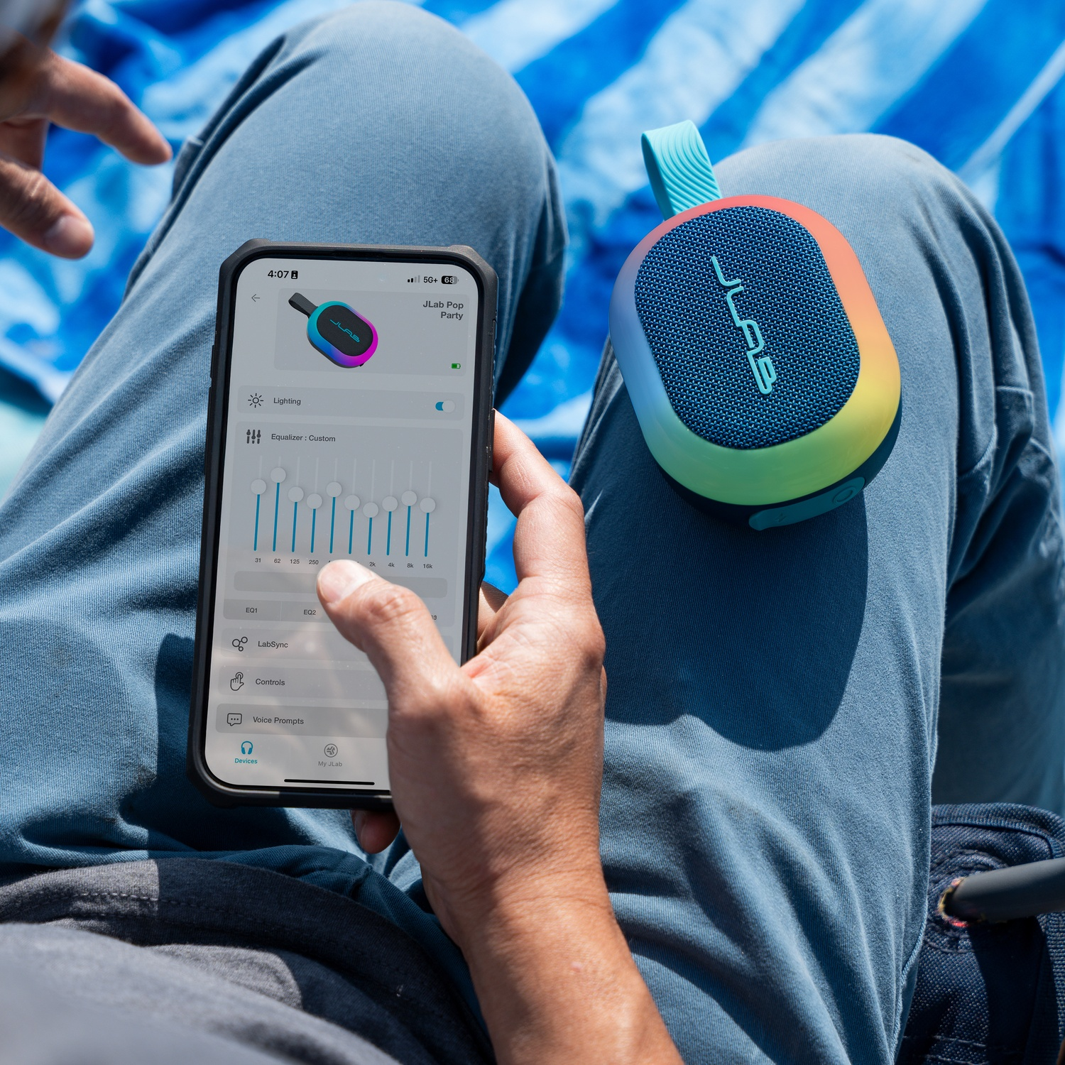 Een persoon gebruikt een telefoon om een JLab Pop Party-luidspreker te bedienen. De luidspreker ligt op de schoot van de persoon. De scène is buiten.