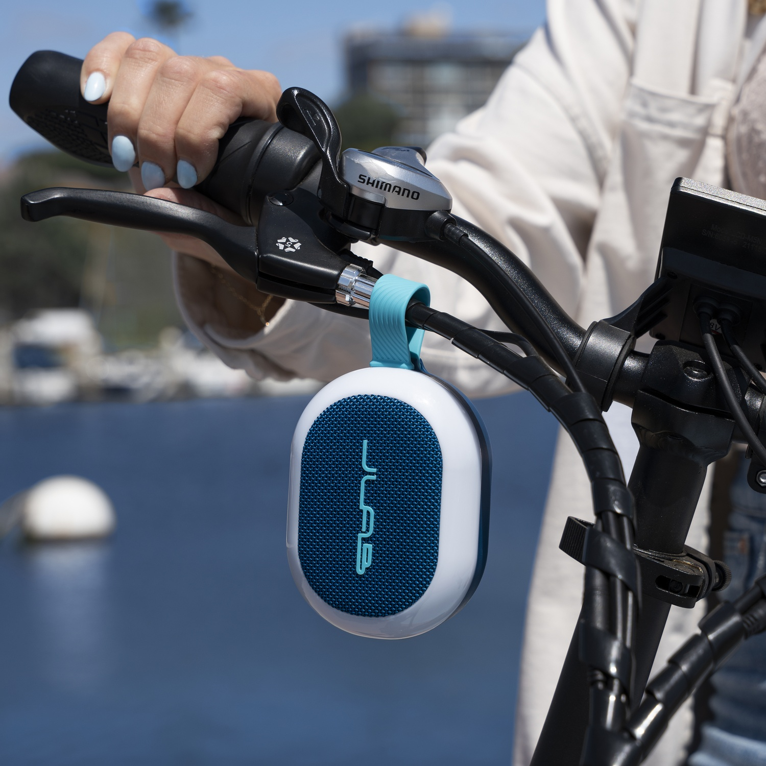 Een persoon houdt het stuur van een fiets vast met een luidspreker eraan. De luidspreker is wit met een blauw gaas. Achtergrond heeft water en gebouwen.