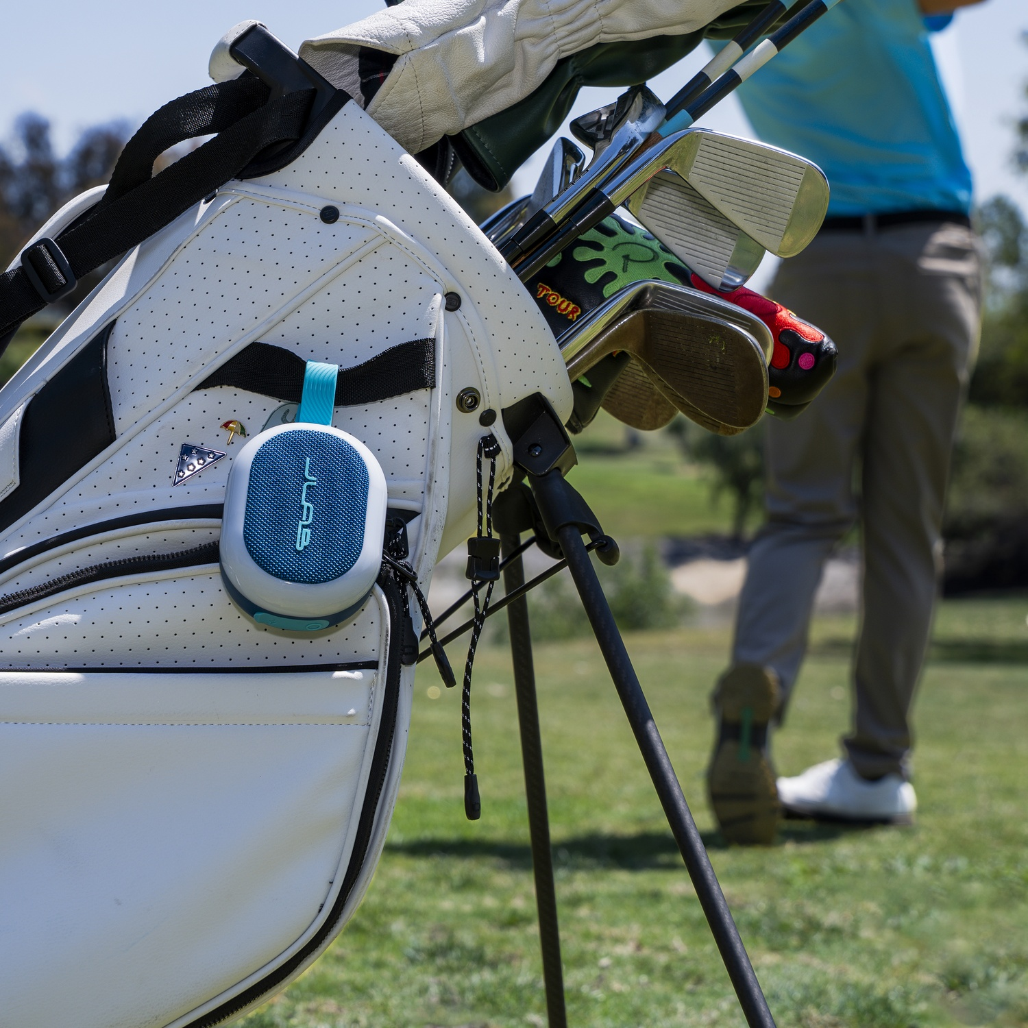 Een golftas met clubs en een luidspreker, een golfer op de achtergrond op een groen veld onder een heldere hemel.