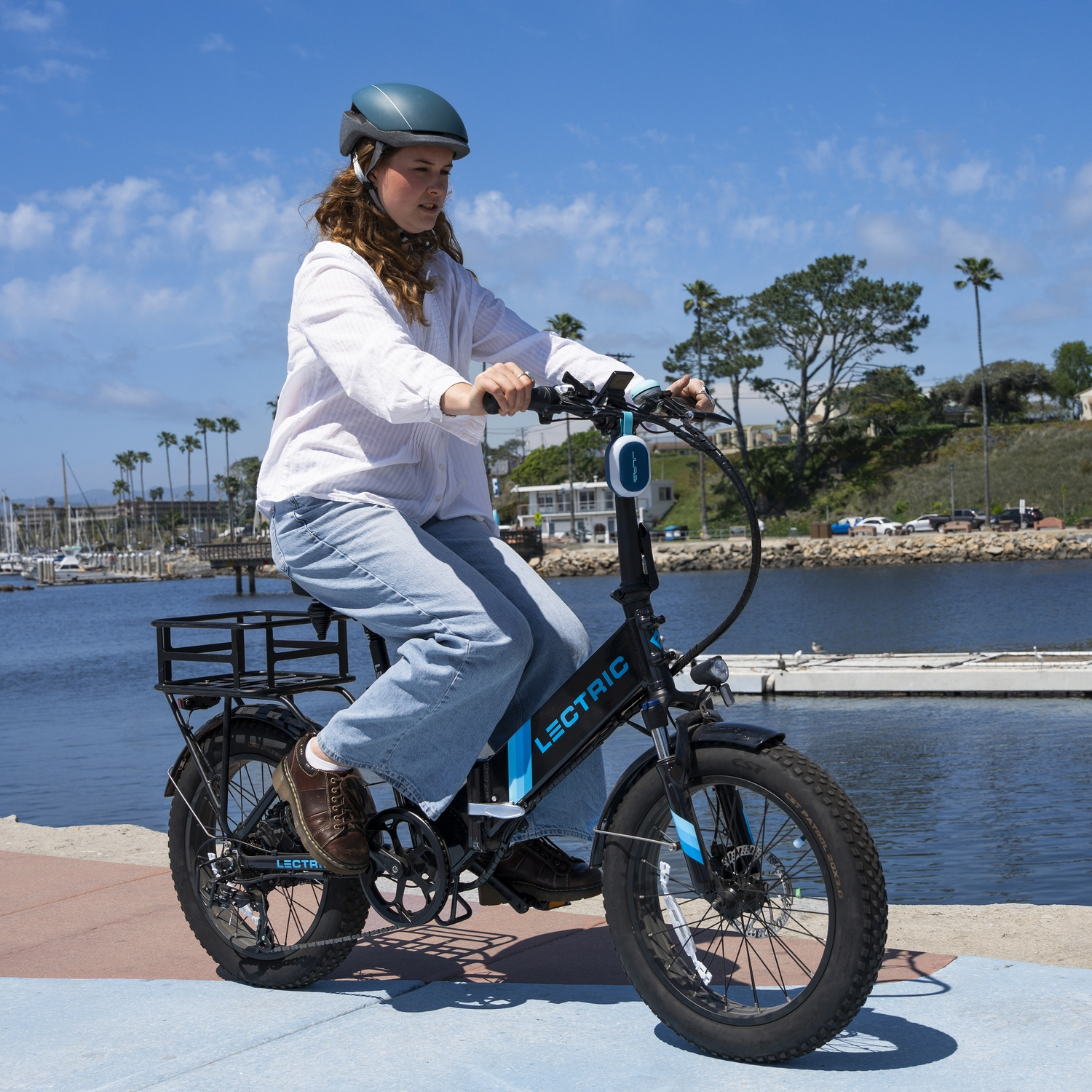 Een vrouw rijdt op een zwarte Lectric e-bike bij een haven. Ze draagt een helm en jeans. Blauwe lucht en palmbomen op de achtergrond.