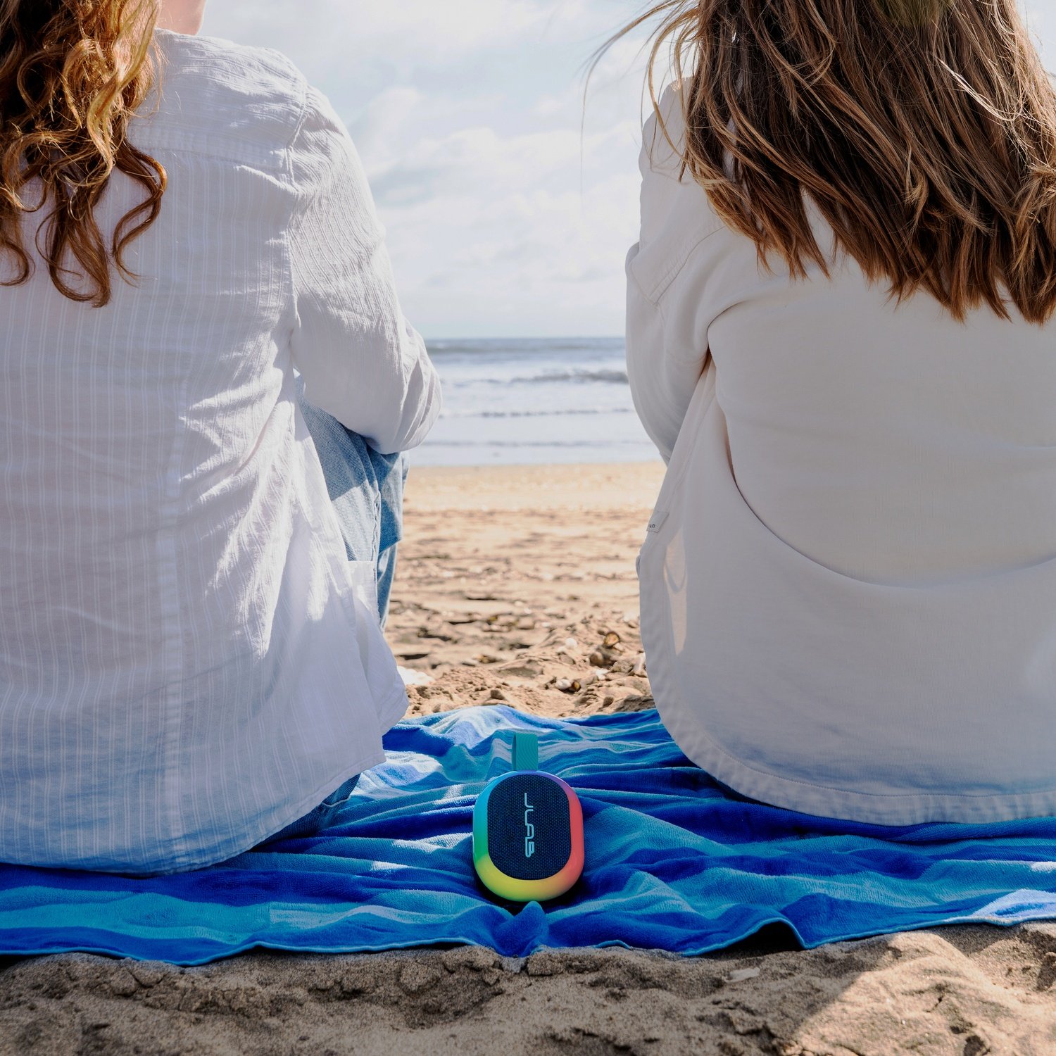 Twee mensen zitten op het strand met uitzicht op de oceaan. Een blauwe handdoek en een kleurrijke luidspreker op de voorgrond. Golven en lucht op de achtergrond.