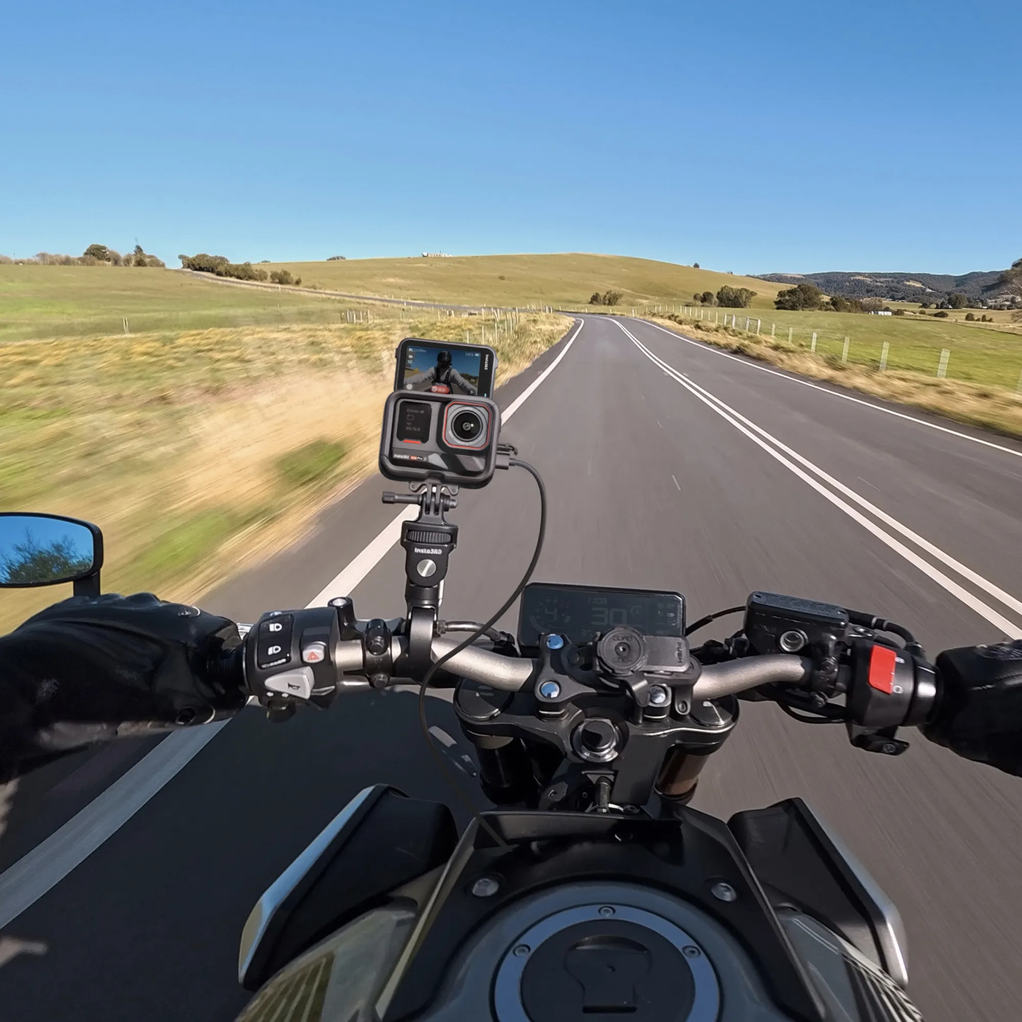 Motorradperspektive. Hände des Fahrers am Lenker, Straße voraus. Kamera montiert. Grüne Felder, Hügel und blauer Himmel im Hintergrund.