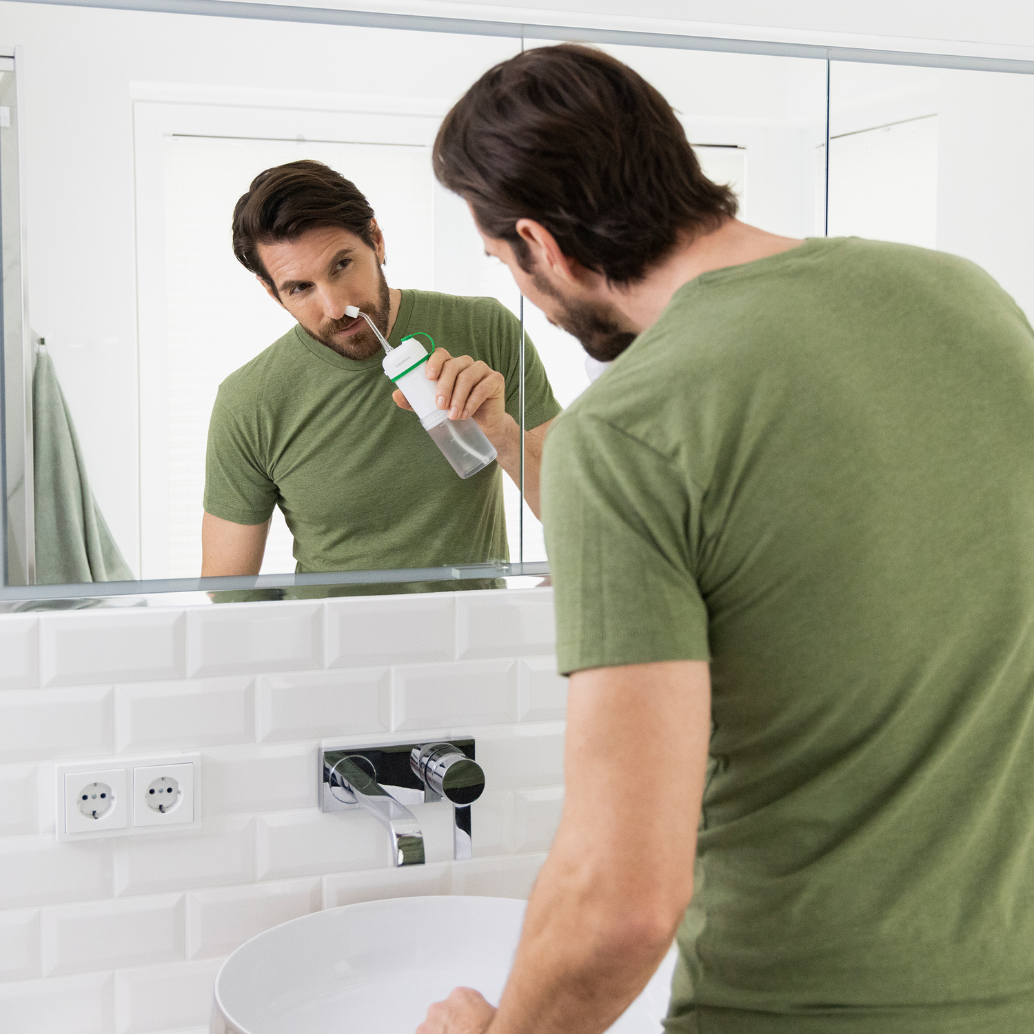 Een man in een groen shirt gebruikt een neusspoeling voor een spiegel in een badkamer. Witte wastafel, tegels en een handdoek zijn zichtbaar.