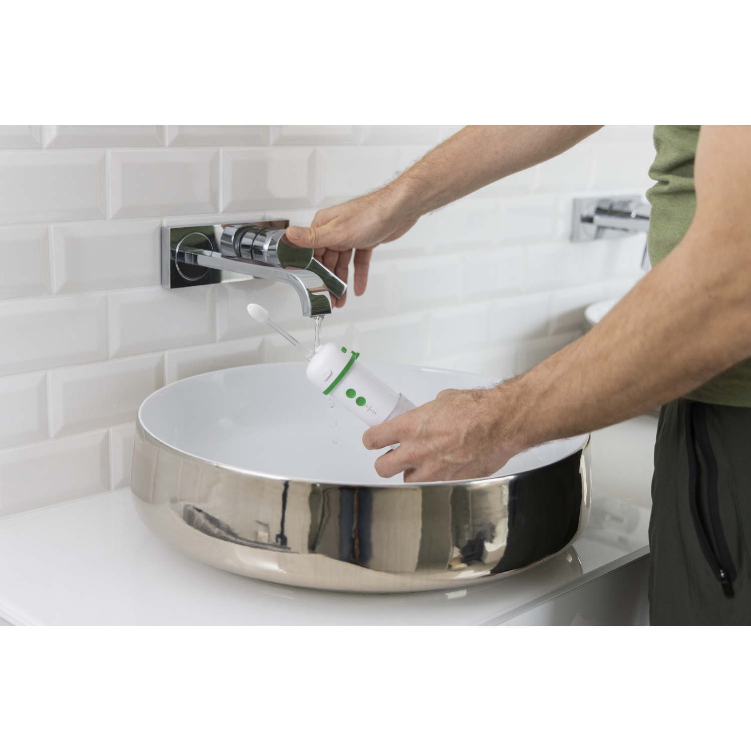 Een persoon vult een witte en groene monddouche met water uit een chromen kraan boven een wastafel. De wastafel is zilver.