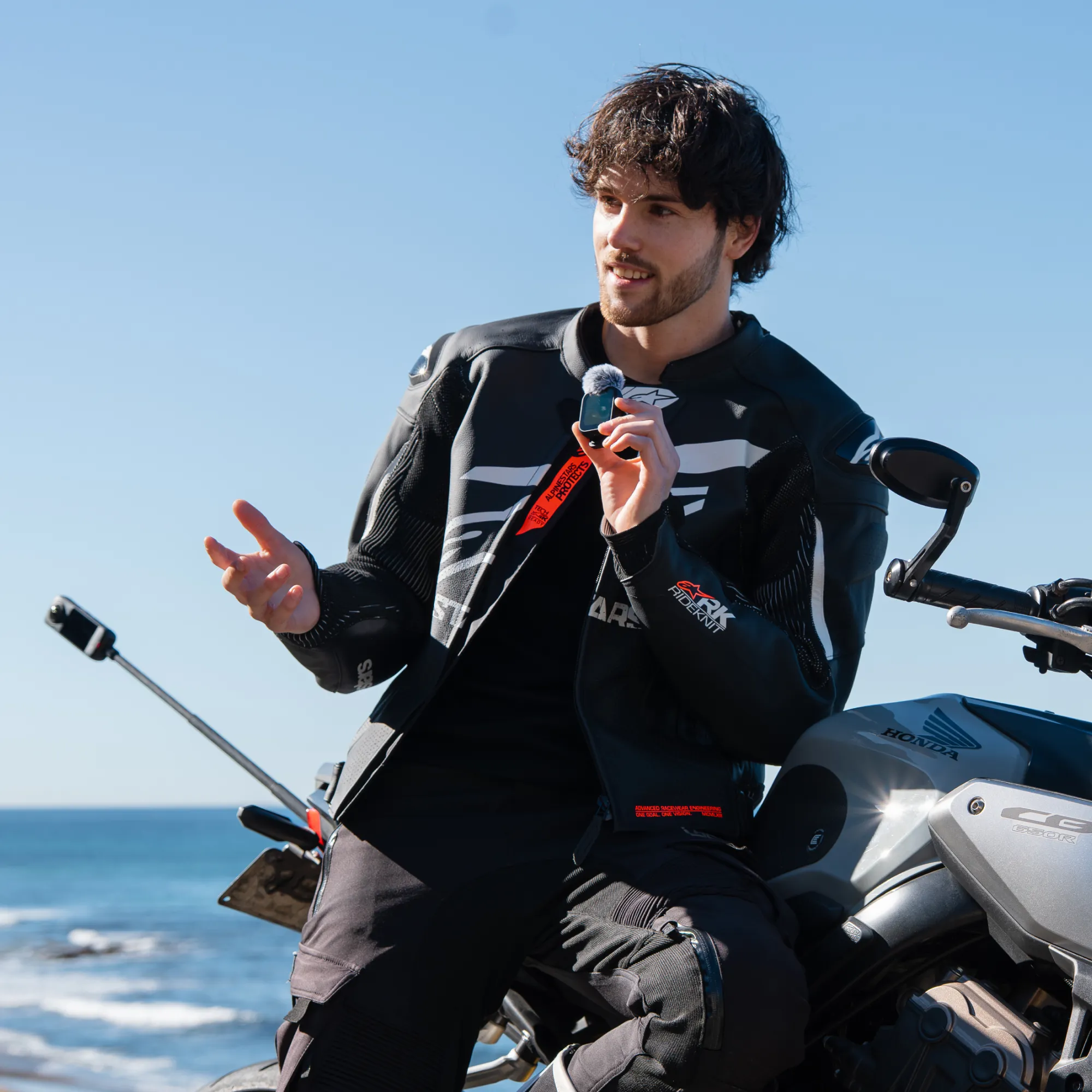 Mann in schwarzer Motorradkleidung mit Mikrofon, lehnt an Honda-Motorrad, Meer im Hintergrund.