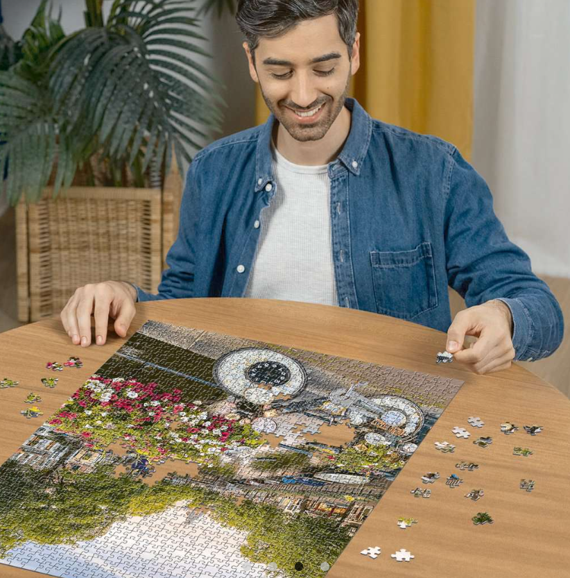 Ein Mann lächelt, während er ein Puzzle auf einem Holztisch zusammensetzt. Das Puzzle zeigt eine Stadtszene mit Blumen, Wasser und Gebäuden.