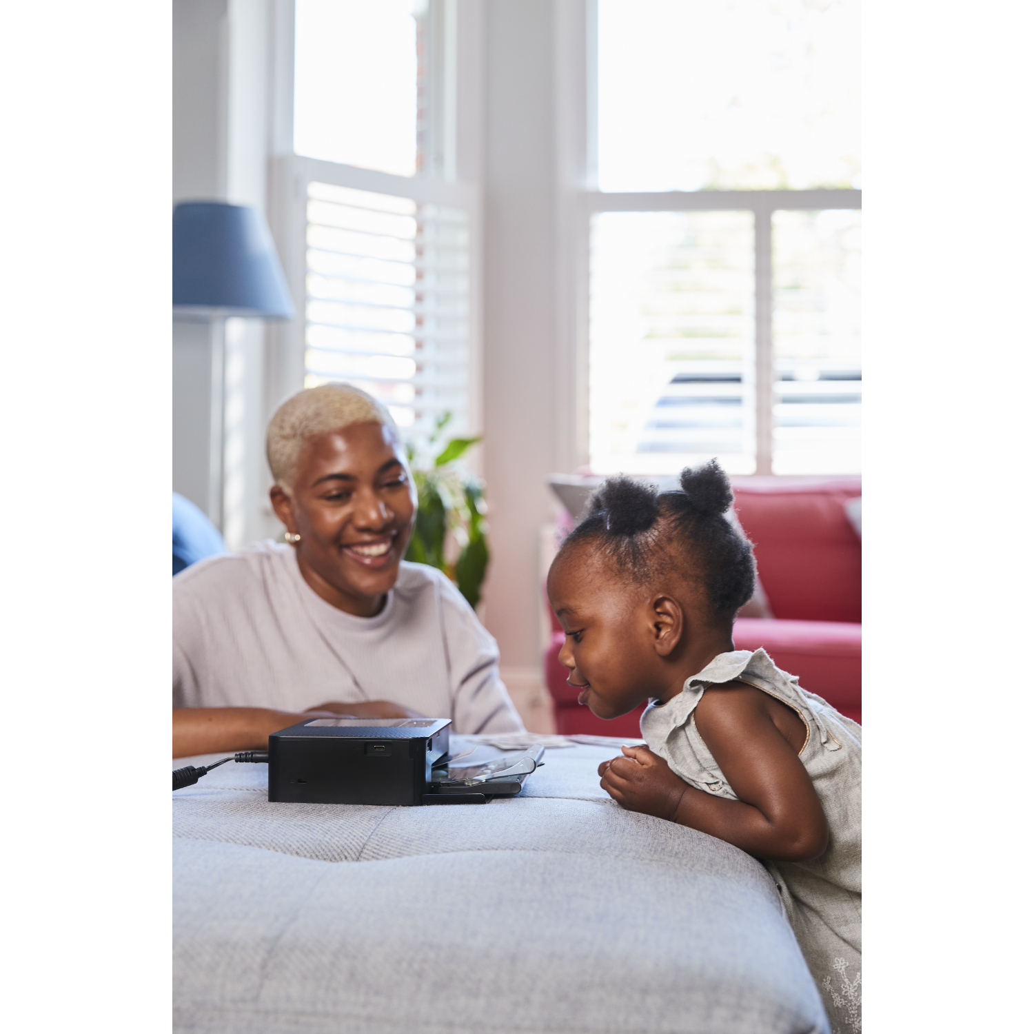 A woman and a child are looking at a black device on a gray surface. The woman is smiling, the child is focused.
