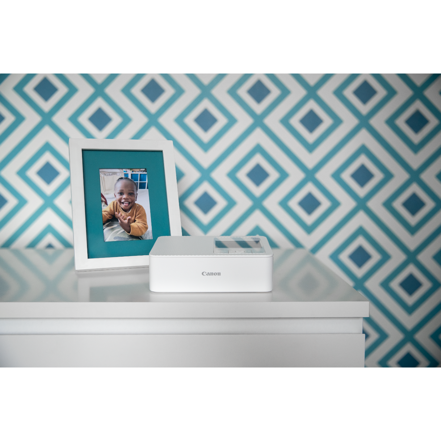 Een witte Canon-printer en een ingelijste foto van een lachend kind staan op een wit oppervlak. De achtergrond heeft een ruitpatroon.