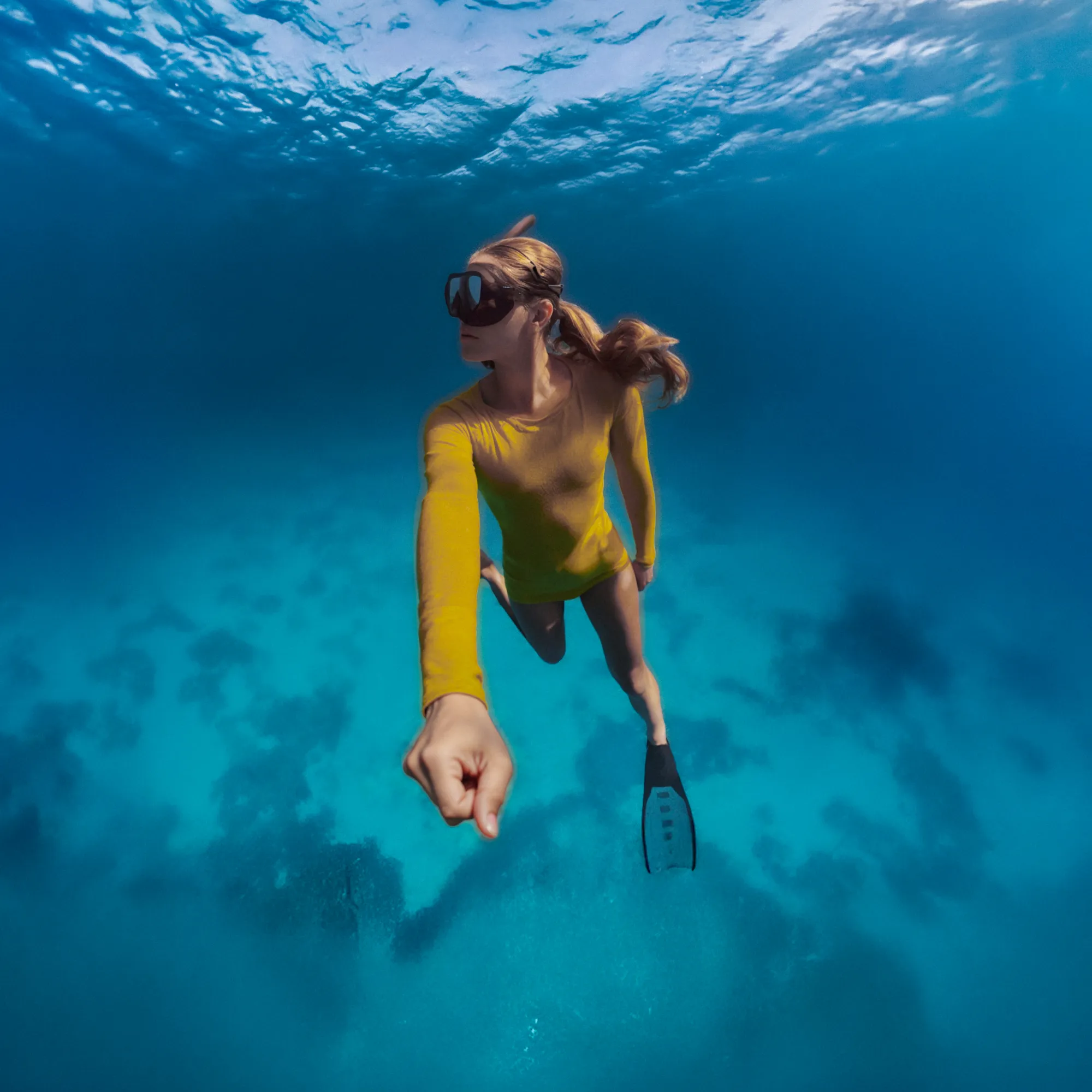 Eine Frau taucht unter Wasser, trägt einen gelben Anzug und eine Maske. Das Wasser ist blau. Sie schwimmt mit Flossen.