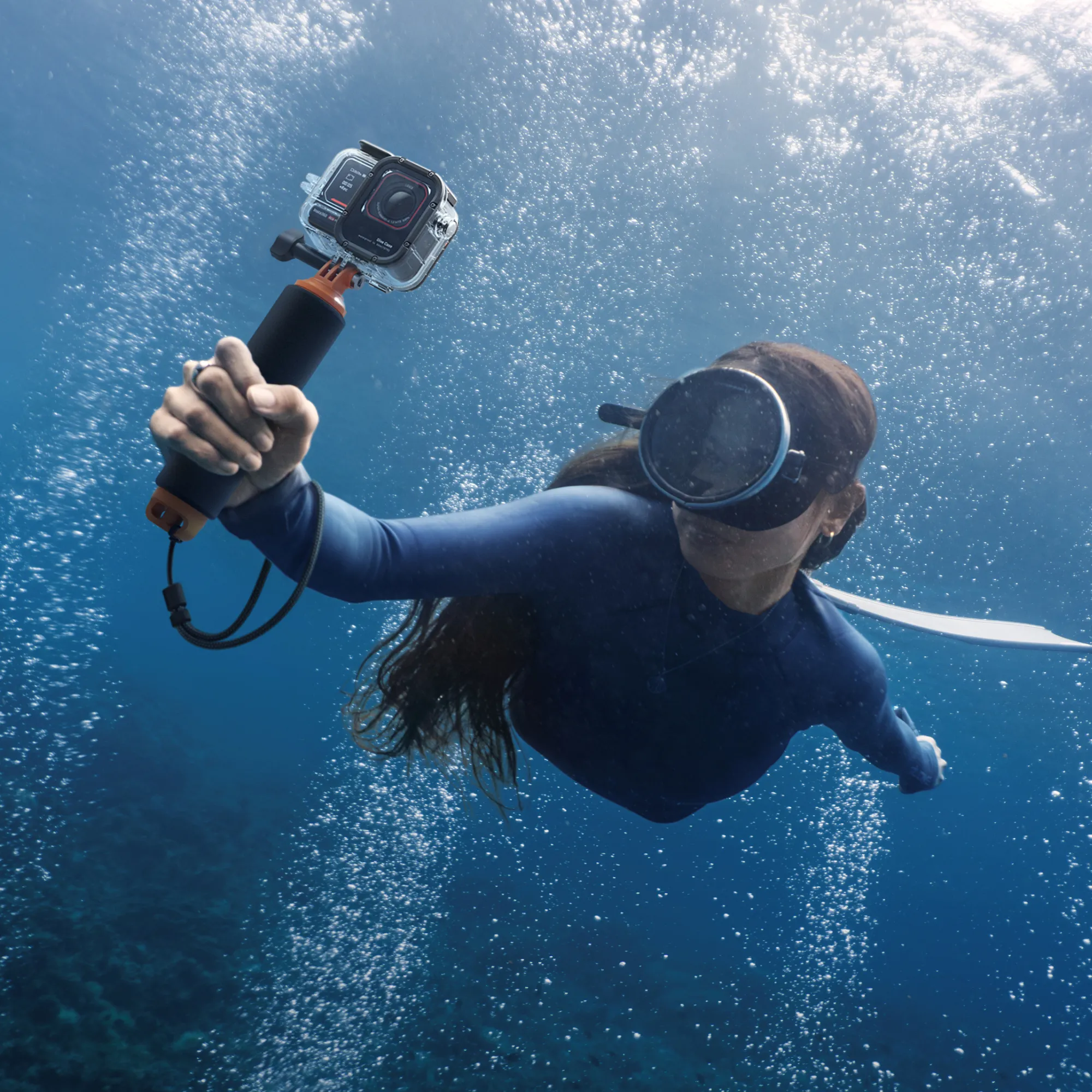 Eine Person unter Wasser hält eine Kamera. Blasen und Sonnenlicht sind im blauen Wasser sichtbar.