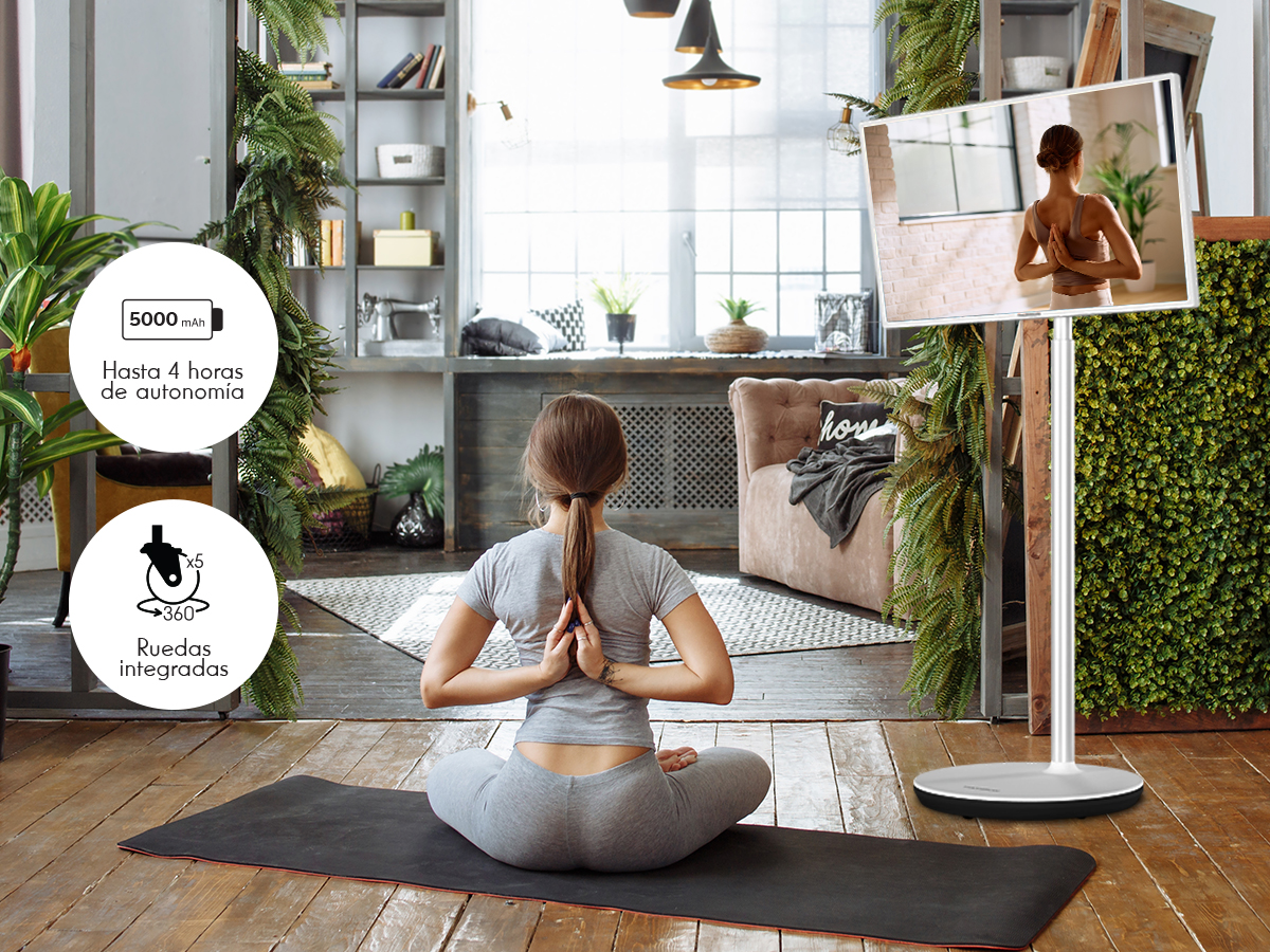 Mujer en pose de yoga, manos atrás, frente a TV. Habitación con plantas, suelo de madera. TV muestra a una mujer en yoga.