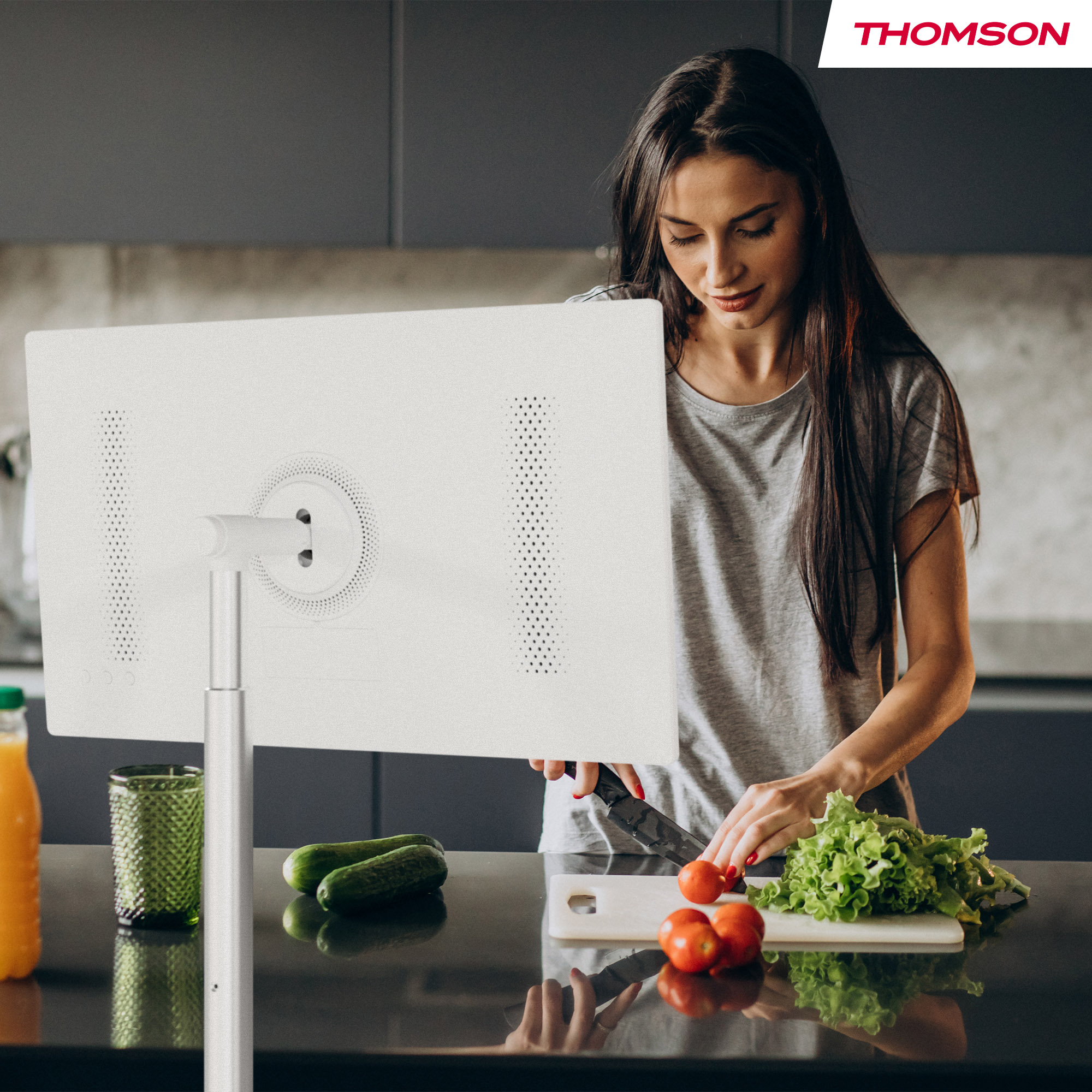Mujer en la cocina cortando tomates. Monitor blanco en primer plano. Pepinos y jugo en la encimera.
