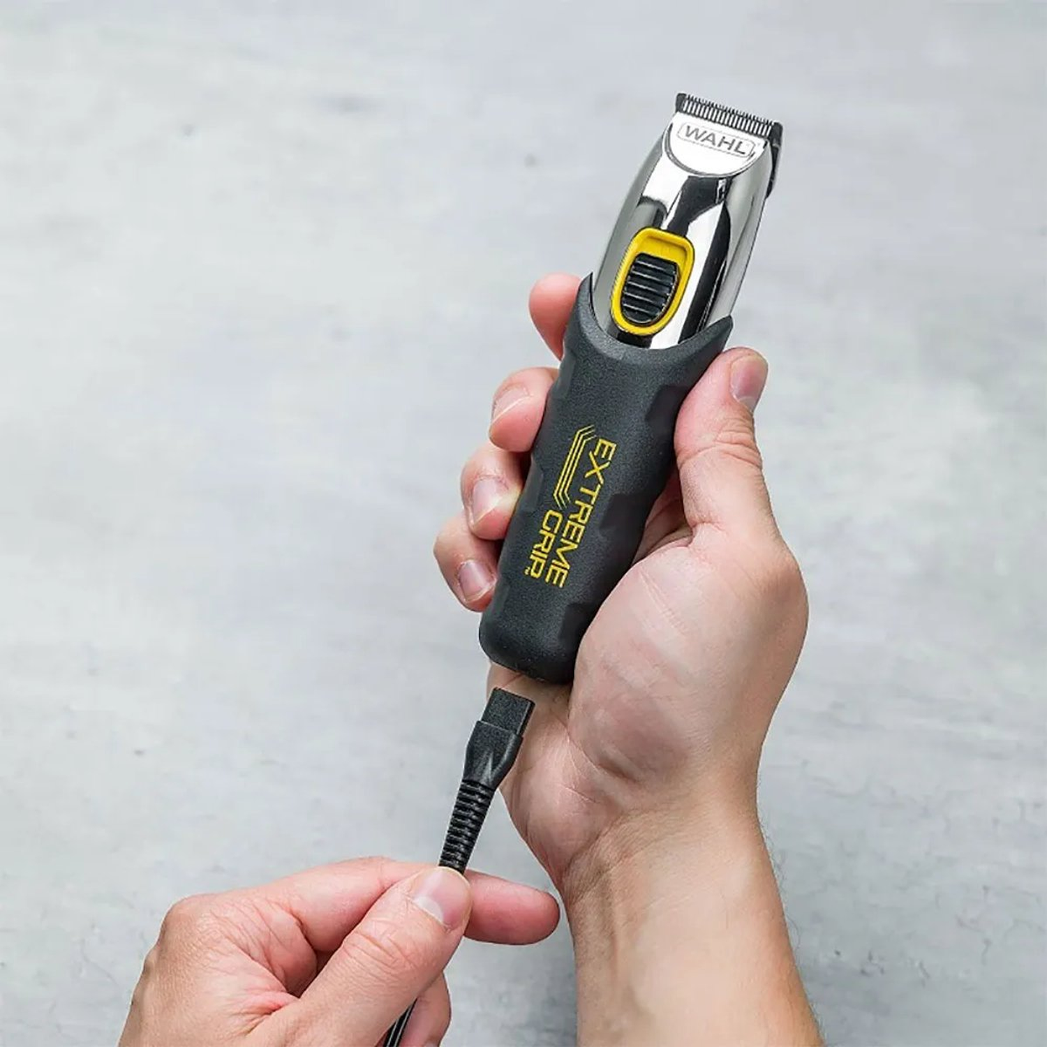 A person holds a black and silver Wahl hair clipper with a black cord. The clipper is held against a gray background.