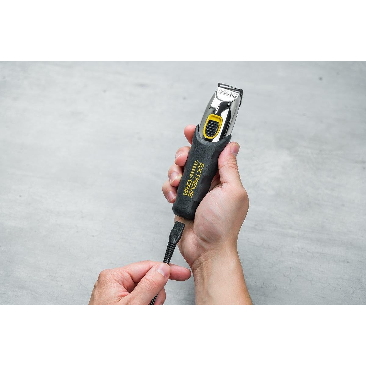 A person holds a Wahl hair clipper with a yellow and black grip. The clipper is silver. The person is holding the cord.