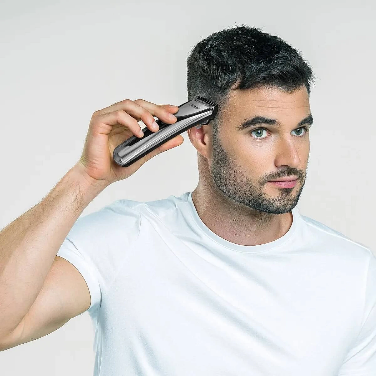 A man in a white t-shirt shaves his hair with an electric trimmer, looking at the camera. The background is white.