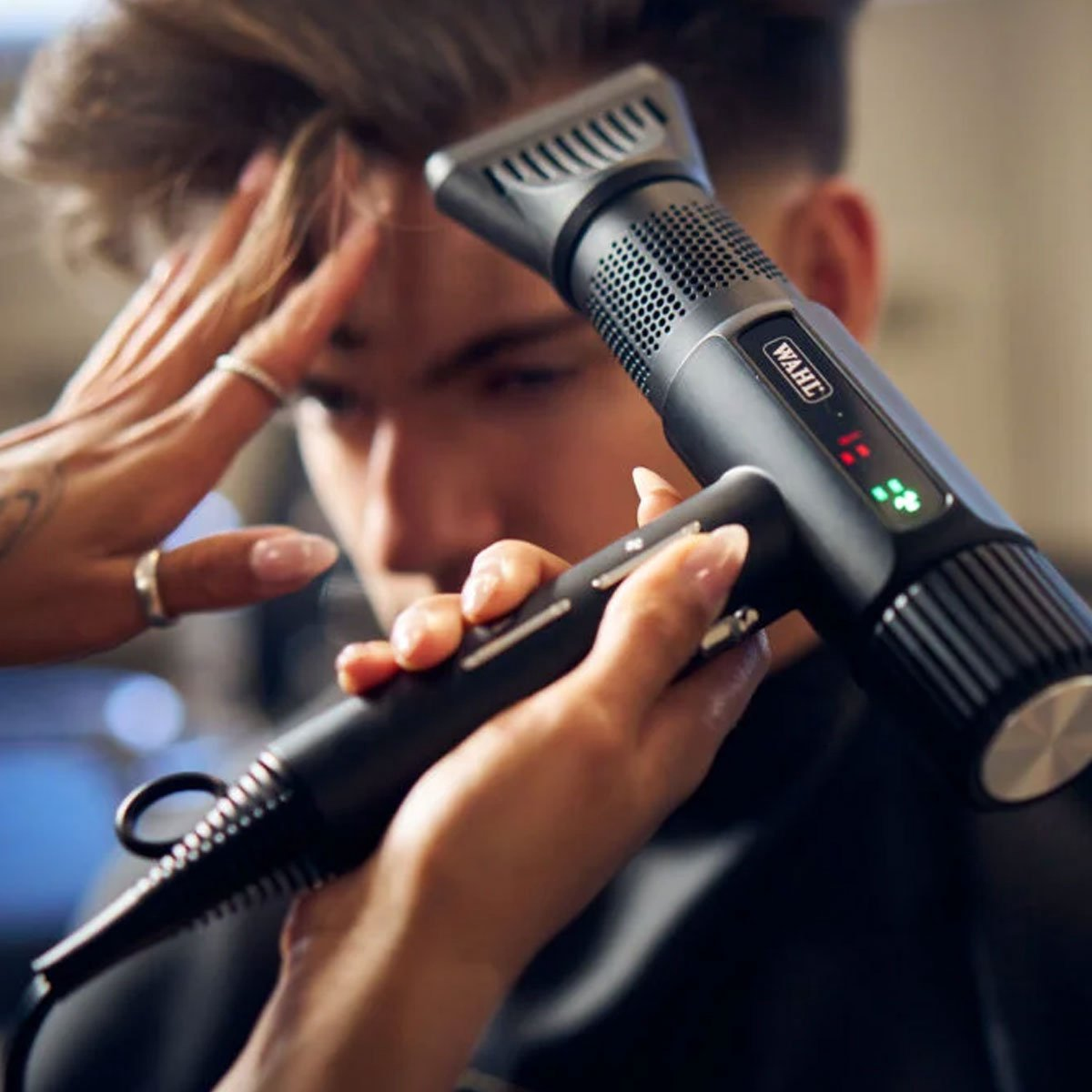 A person is using a black Wahl hair dryer on another person's hair. The dryer is held by two hands. The background is blurred.