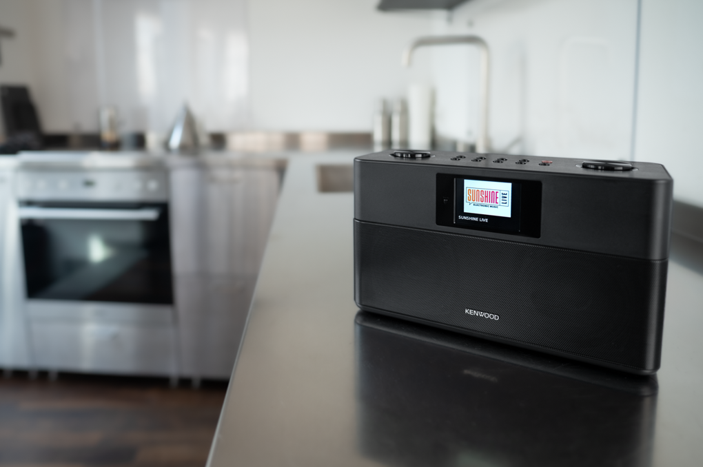 Una radio Kenwood nera con schermo che mostra 'Sunshine Live' è appoggiata su un bancone. Elettrodomestici sfocati sullo sfondo.