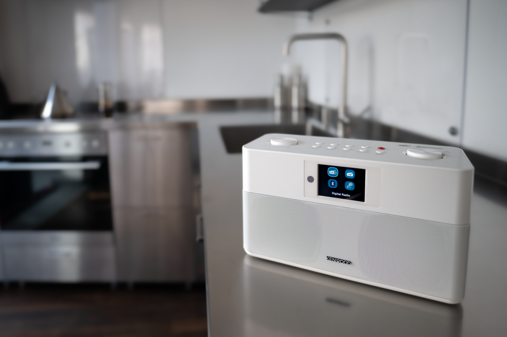 Una radio Kenwood bianca su un bancone in acciaio inossidabile. Lo sfondo della cucina è sfocato.