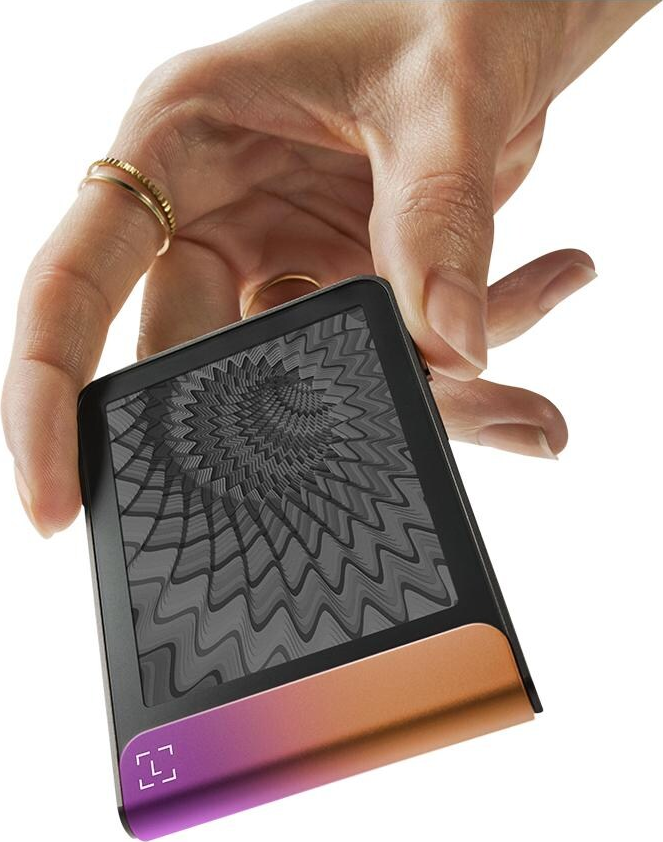 Una mano tiene in mano un dispositivo con uno schermo a motivi. Il dispositivo ha una sfumatura dal viola all'arancione. Anelli d'oro adornano la mano.