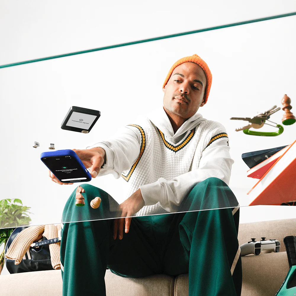 Ein Mann in einer orangefarbenen Mütze und einem weißen Pullover hält ein Telefon. Schachfiguren, Schlüssel und ein Tablet schweben um ihn herum. Er sitzt auf einem Sofa.