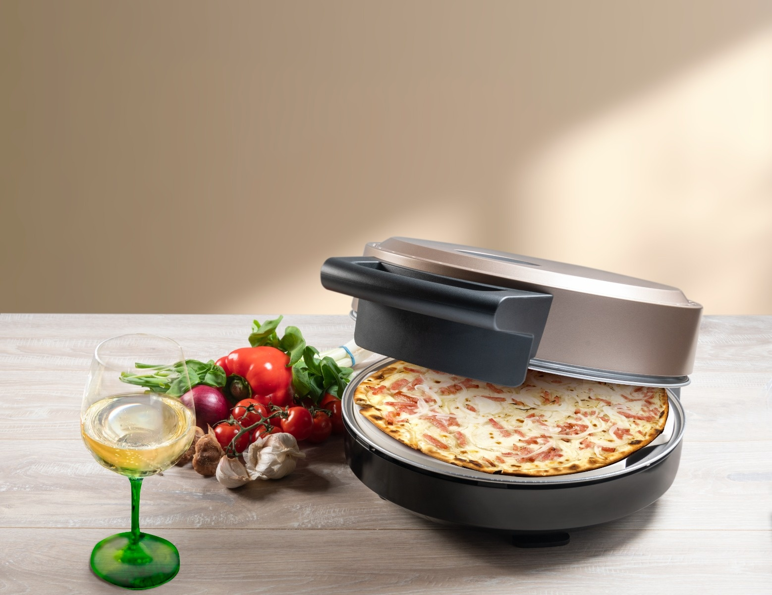 Eine Pizza befindet sich in einem Elektroofen. Daneben stehen ein Weinglas und Gemüse. Der Hintergrund ist eine beige Wand.