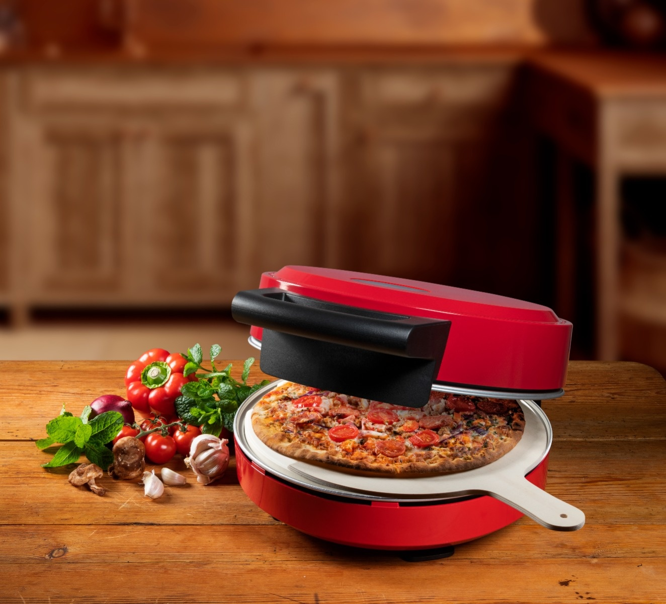 Ein roter Pizzaofen mit einer Pizza darin, umgeben von Gemüse auf einem Holztisch. Der Hintergrund ist eine verschwommene Küche.