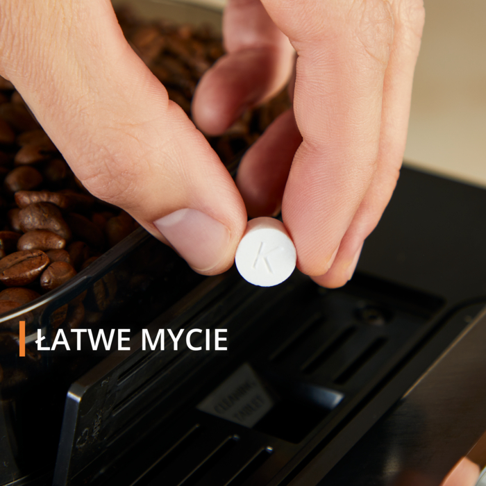 Dłoń trzyma białą tabletkę z literą 'K' nad pojemnikiem na ziarna kawy. Widoczny jest napis 'ŁATWE MYCIE'.