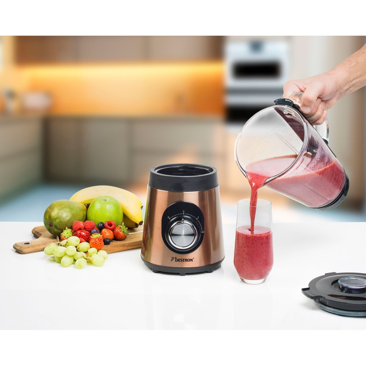 Eine Hand gießt roten Smoothie aus einem Mixer in ein Glas. Früchte wie Bananen, Trauben und Mangos liegen neben dem Mixer.